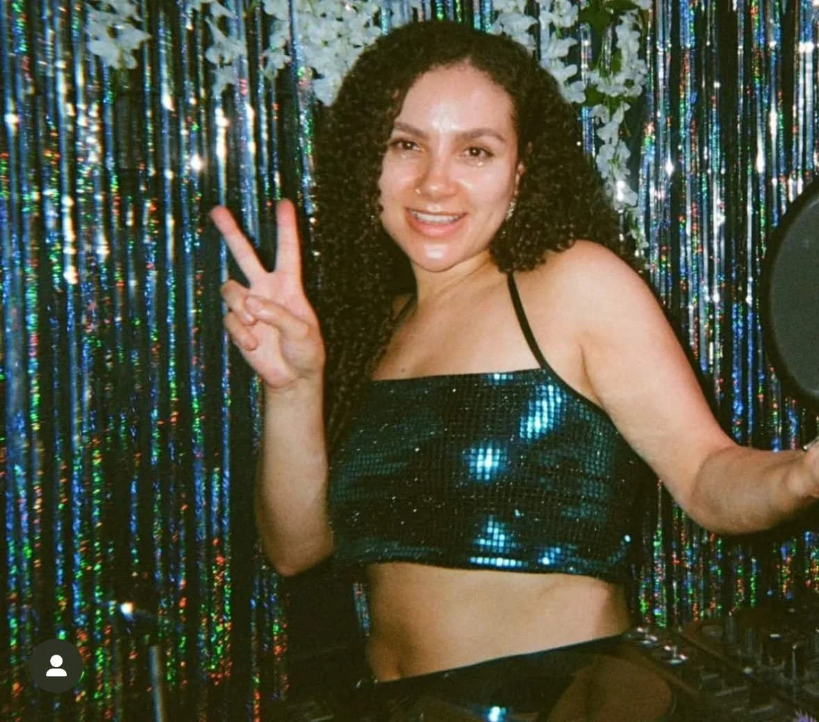 Woman with curly hair making a peace sign, standing in front of a colorful, shiny party backdrop, wearing a sparkly black crop top.