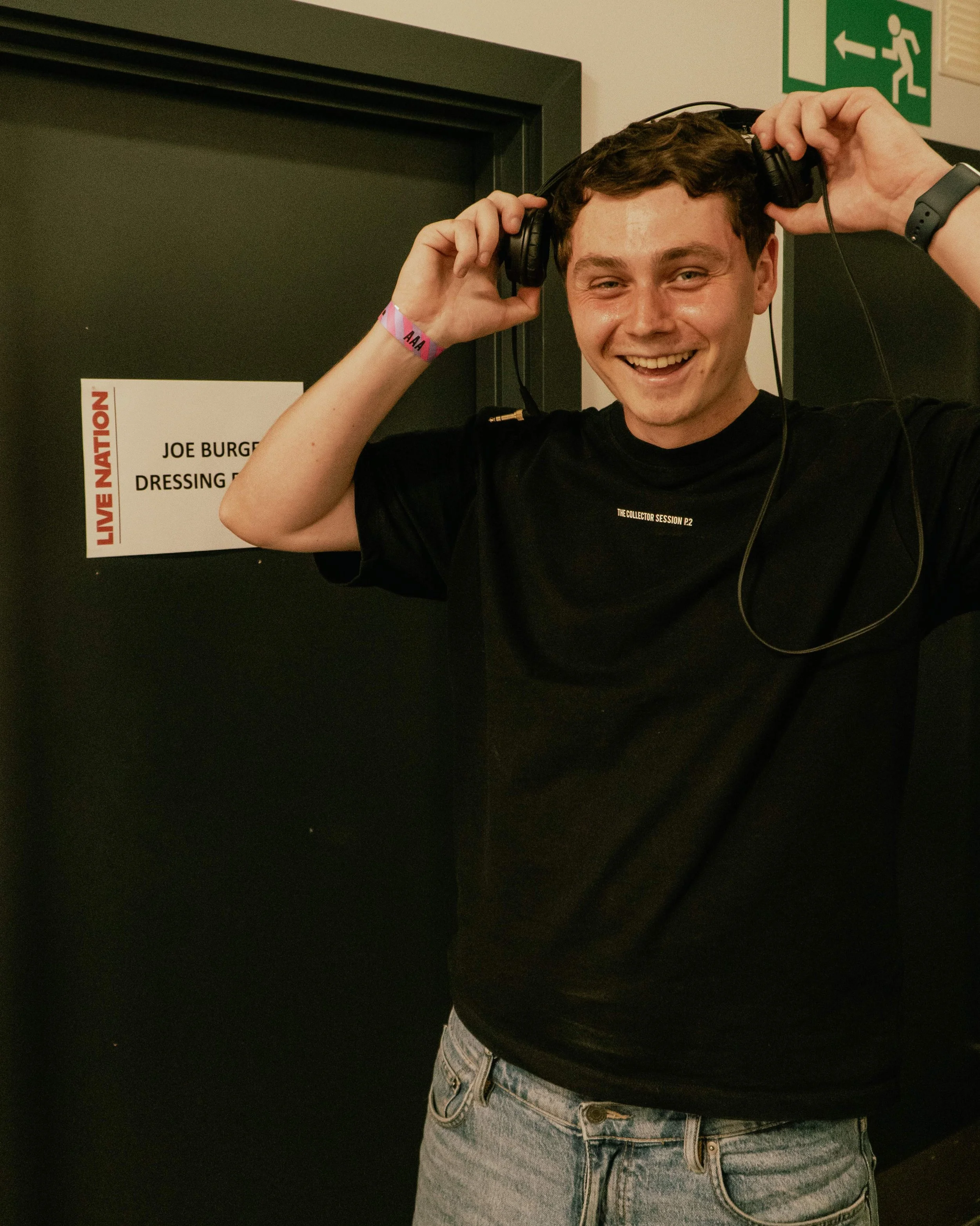 A young man with short brown hair, smiling and holding a pair of headphones on his head, standing in front of a dark door with a sign that says 'Joe Burgess Dressing.' He is wearing a black t-shirt with small text on the chest and a pink wristband on his right wrist. An emergency exit sign is visible in the background.