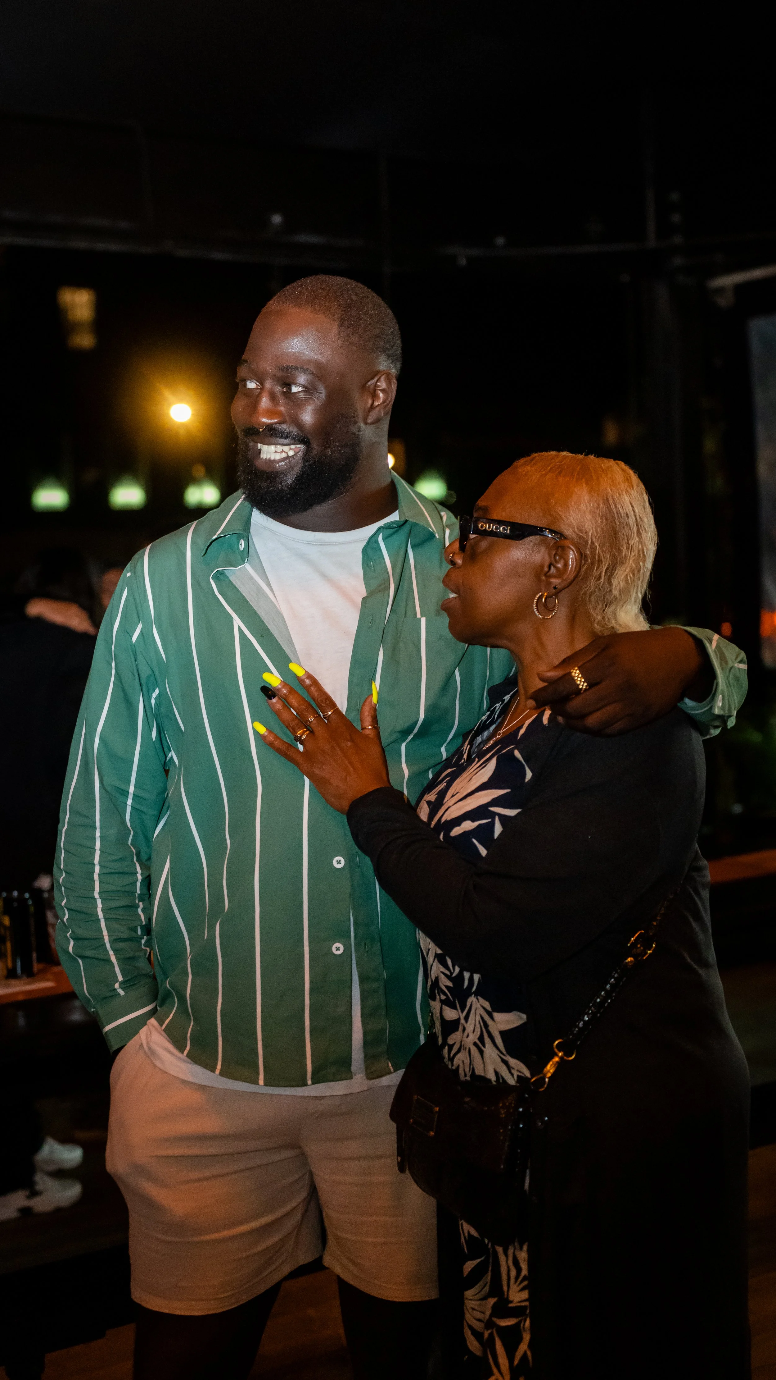 A man with a beard and a woman with short blonde hair wearing glasses are talking and smiling at night in a dimly lit environment.