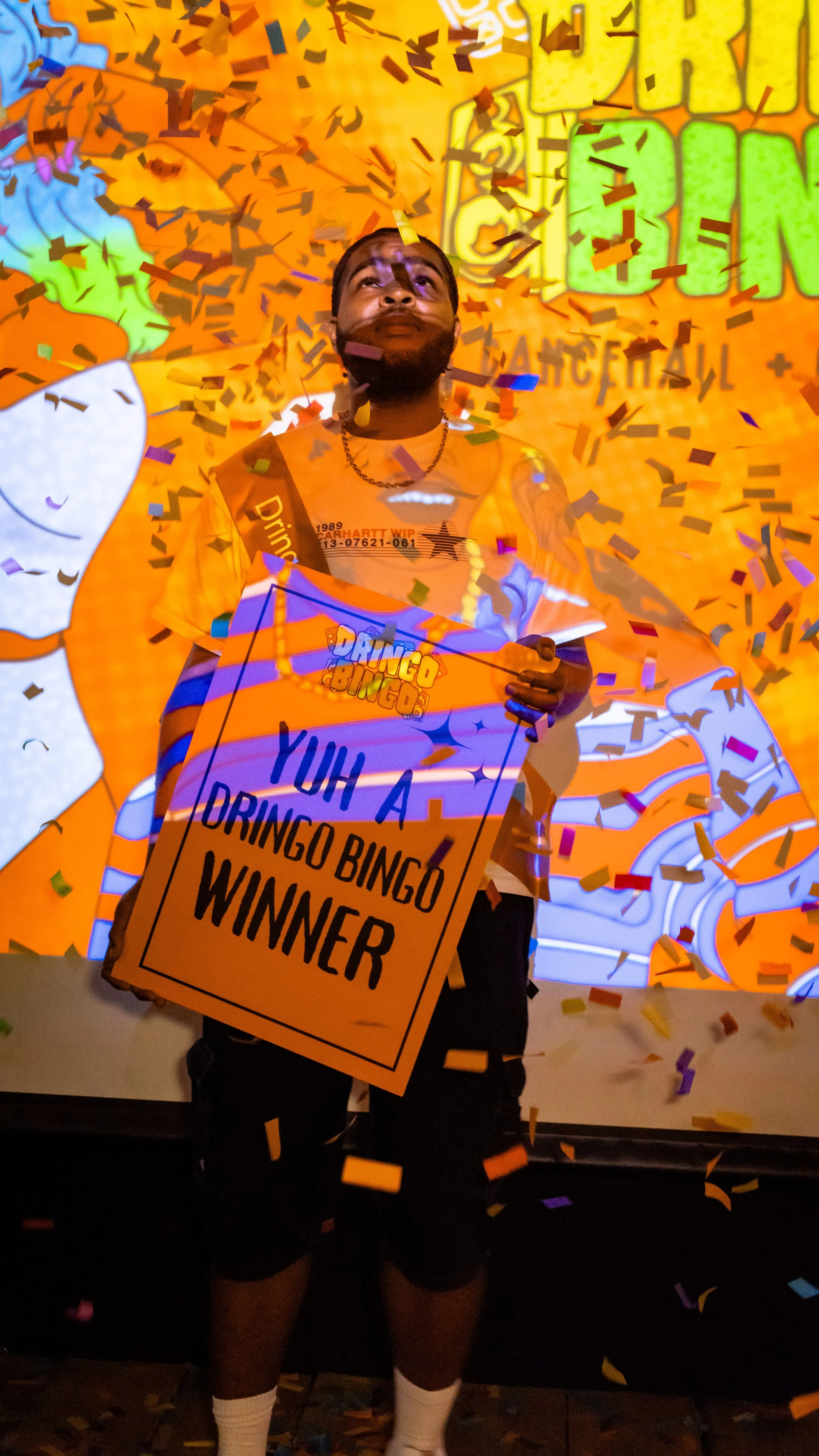 A man holding a sign that says 'Yuh A Dringo Bingo Winner' surrounded by falling confetti at a celebration event.