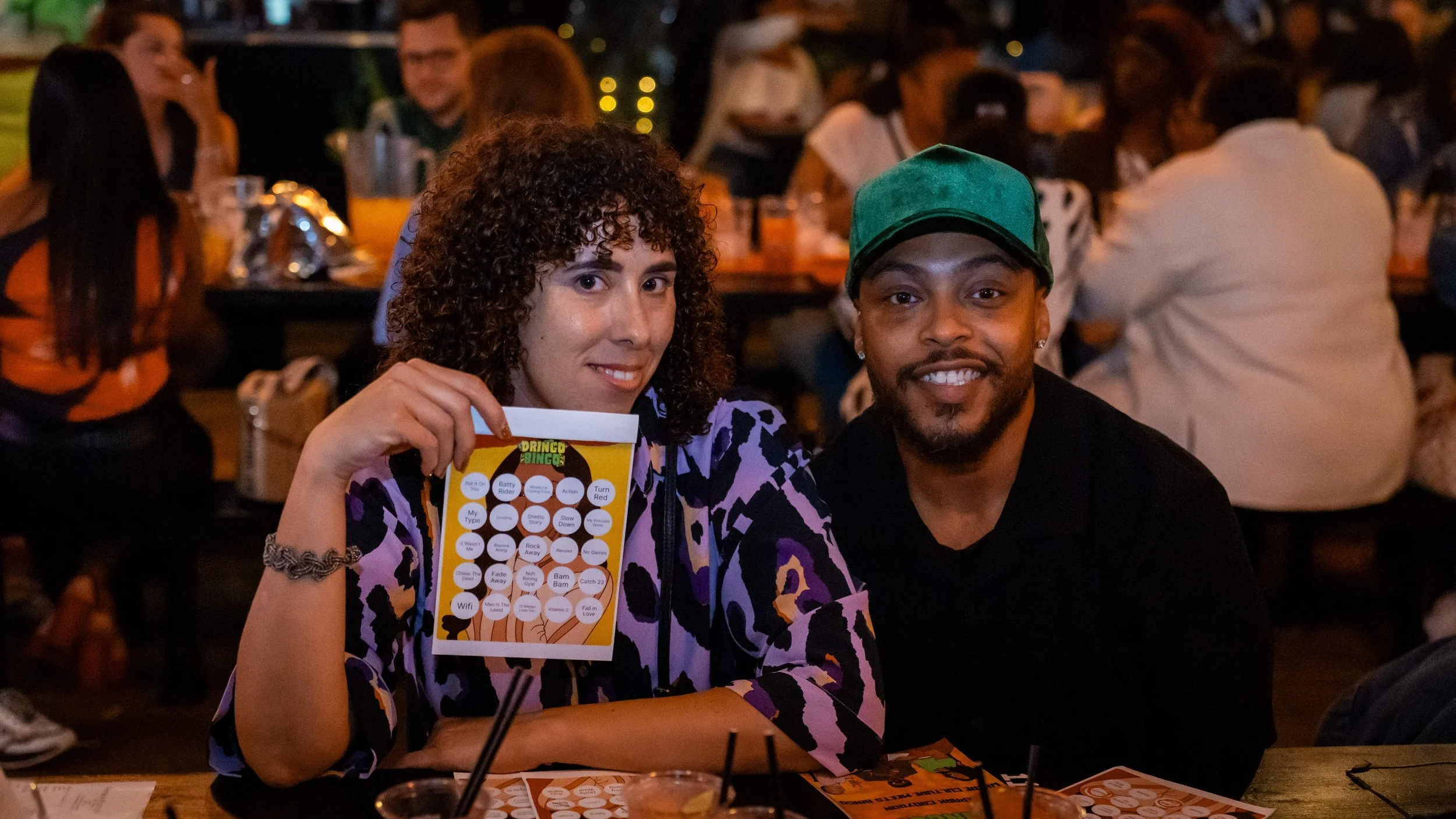 A man and woman sitting at a table together at a social event, smiling at the camera. The woman has curly hair and is holding a bingo card. The man is wearing a green cap and a black shirt. The background shows other people sitting at tables, chatting and enjoying drinks.