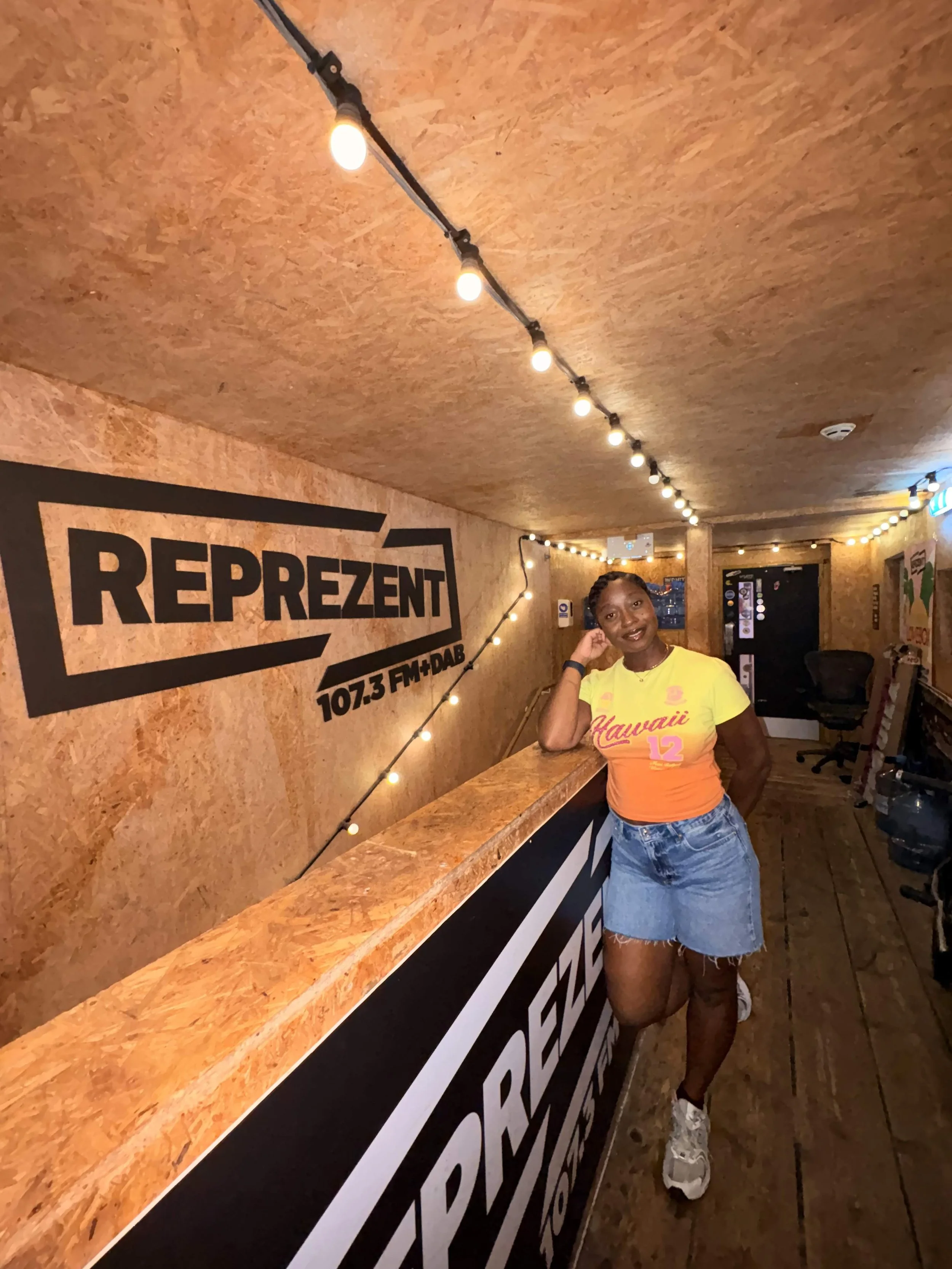 Woman standing next to a wooden radio station counter, wearing a yellow and orange t-shirt with 'Hawaii 12' written on it and denim shorts, inside a studio with string lights and a large sign that reads 'REPREZENT 107.3 FM + DAB'.