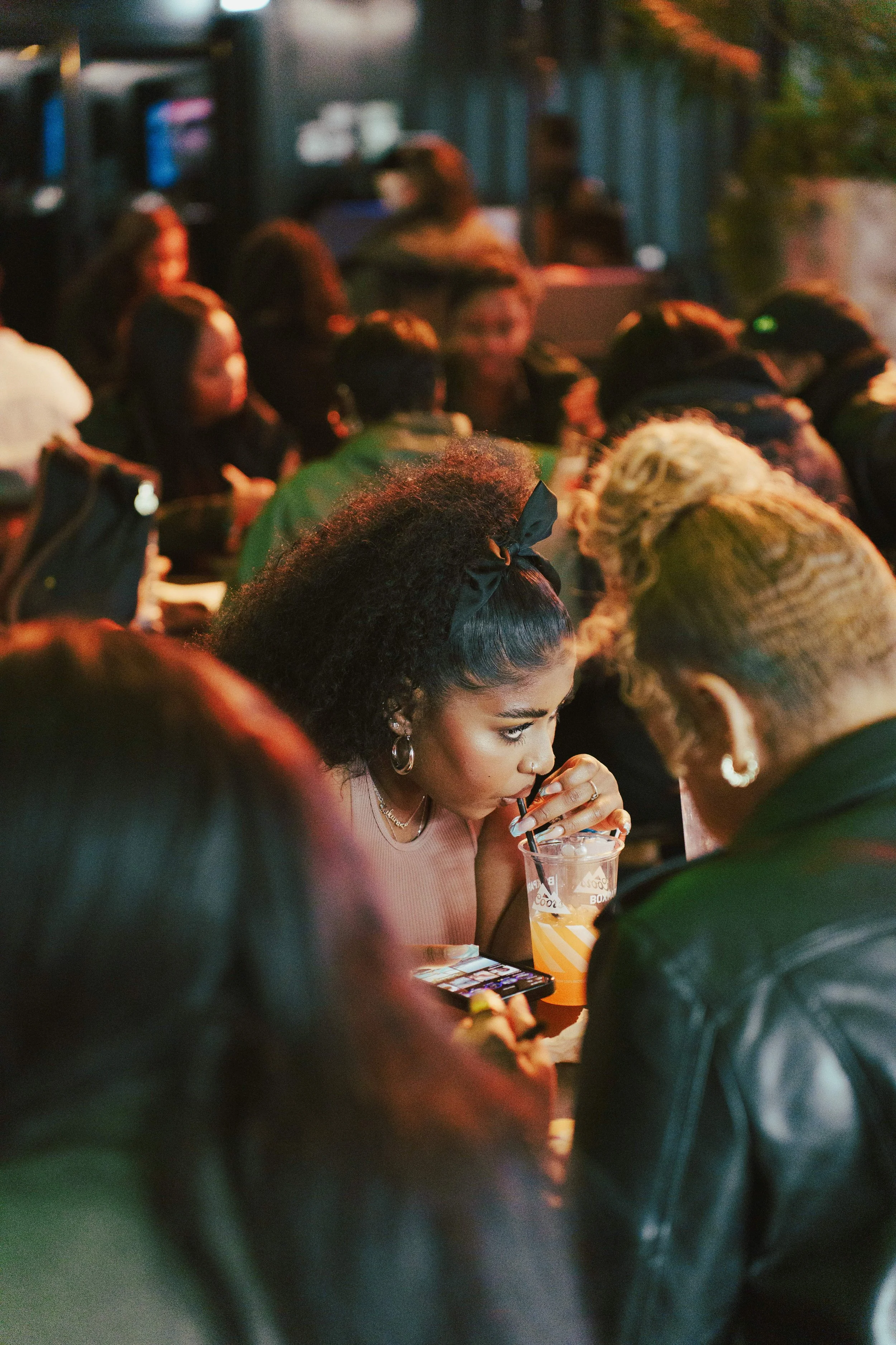 A woman drinking a beverage with a straw at a crowded outdoor event during evening hours.