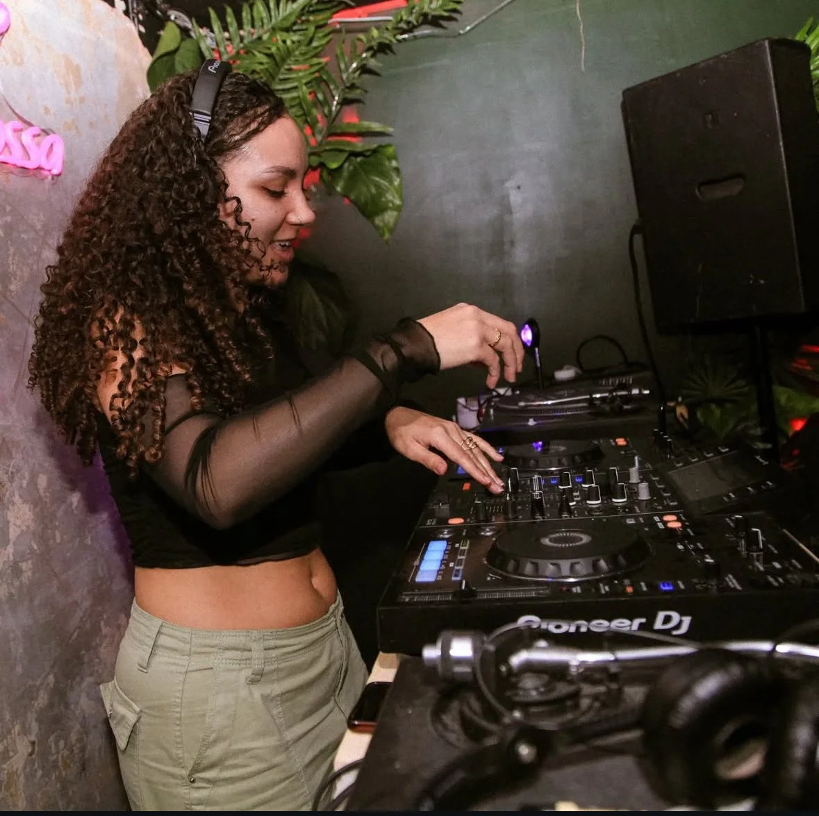 A woman DJ with long curly hair wearing a black sheer top and beige pants, operating a Pioneer DJ controller at a nightclub or party.
