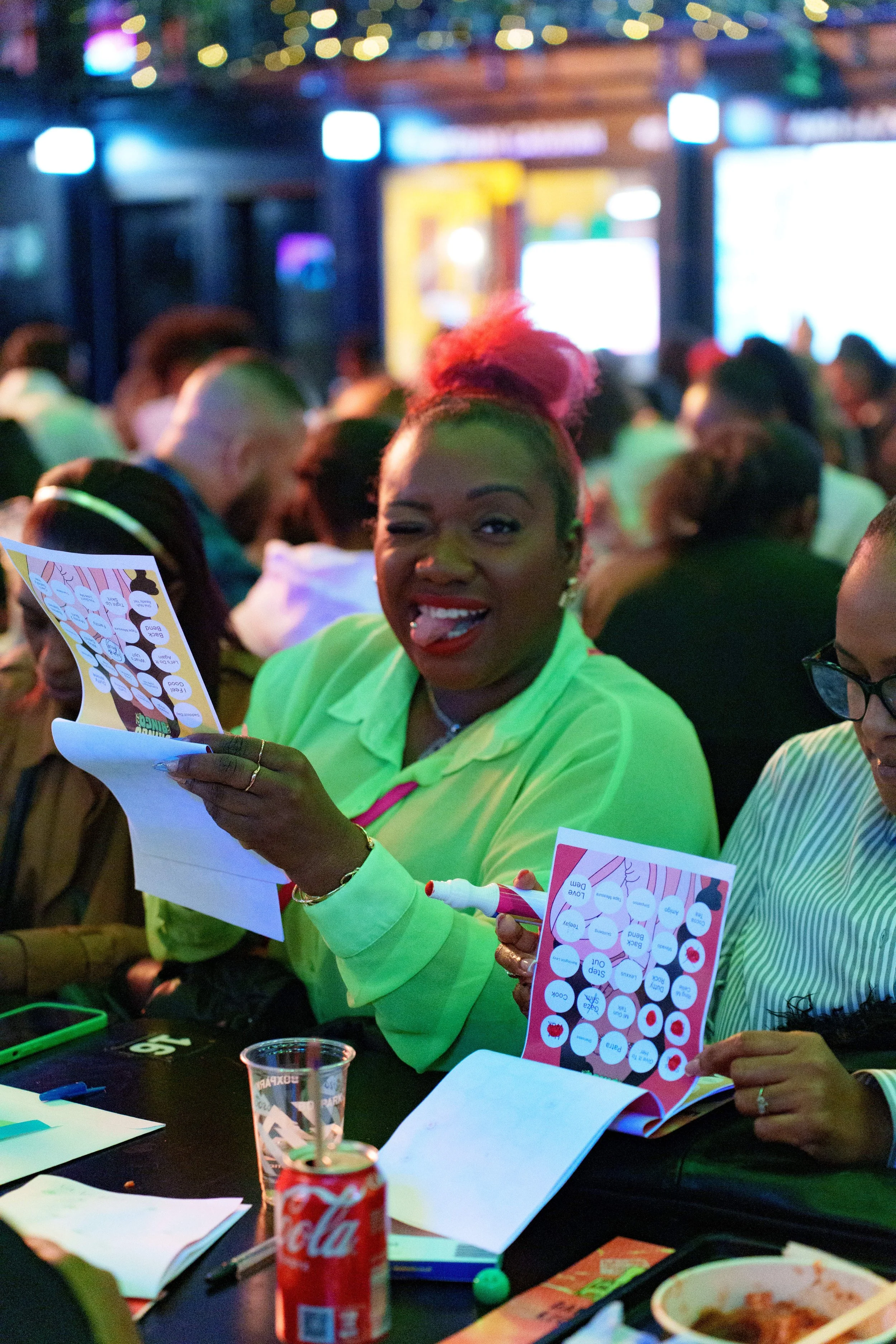 A woman with pink hair styled in a high bun, wearing a bright green shirt, is sitting at a table filled with papers, a pen, a can of Coca-Cola, and a clear plastic cup. She is winking, sticking out her tongue, and holding a bingo card while pointing