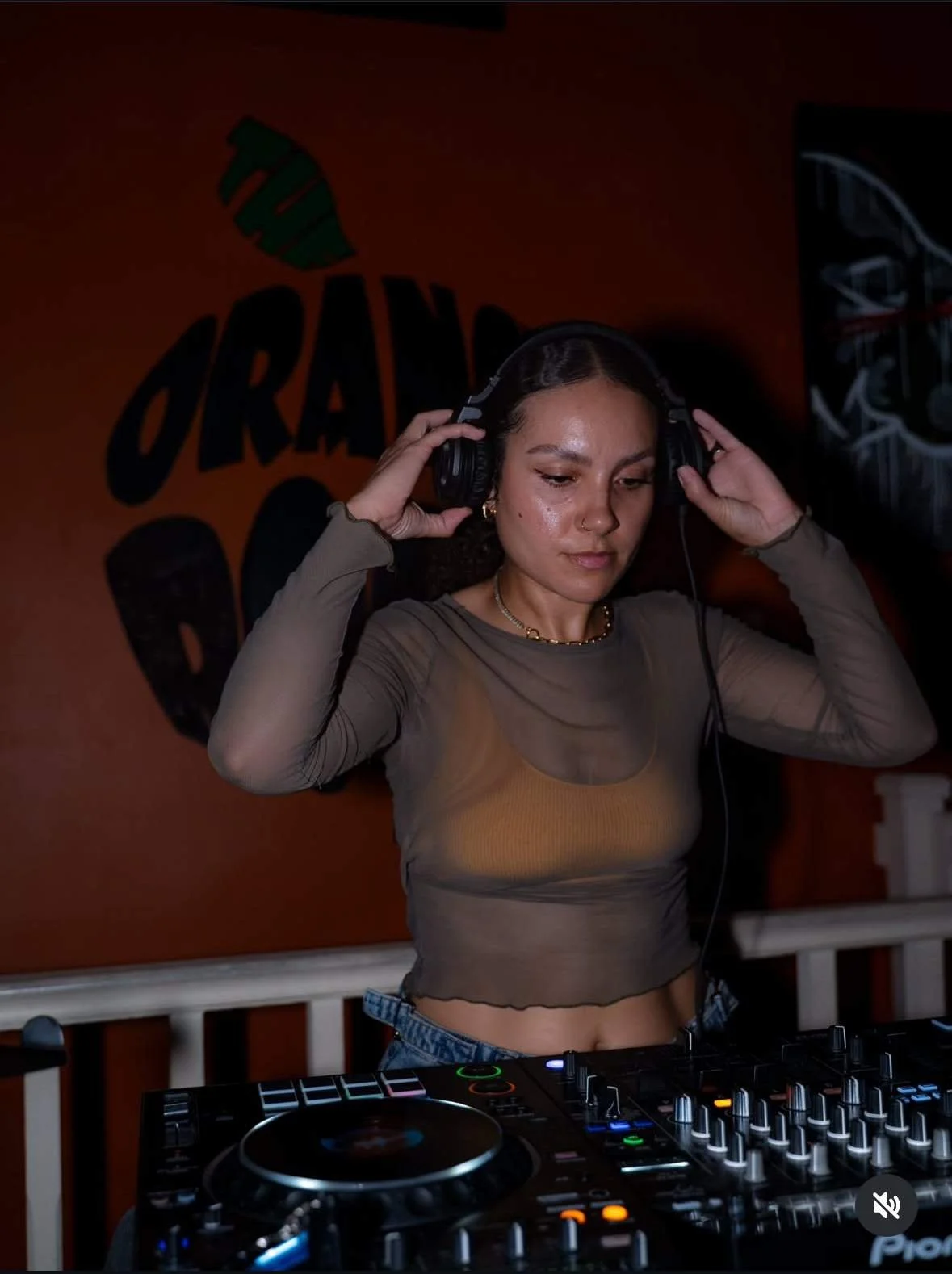 A woman with curly hair wearing a sheer long-sleeve top and jeans, is DJing with headphones on at a nightclub or party.