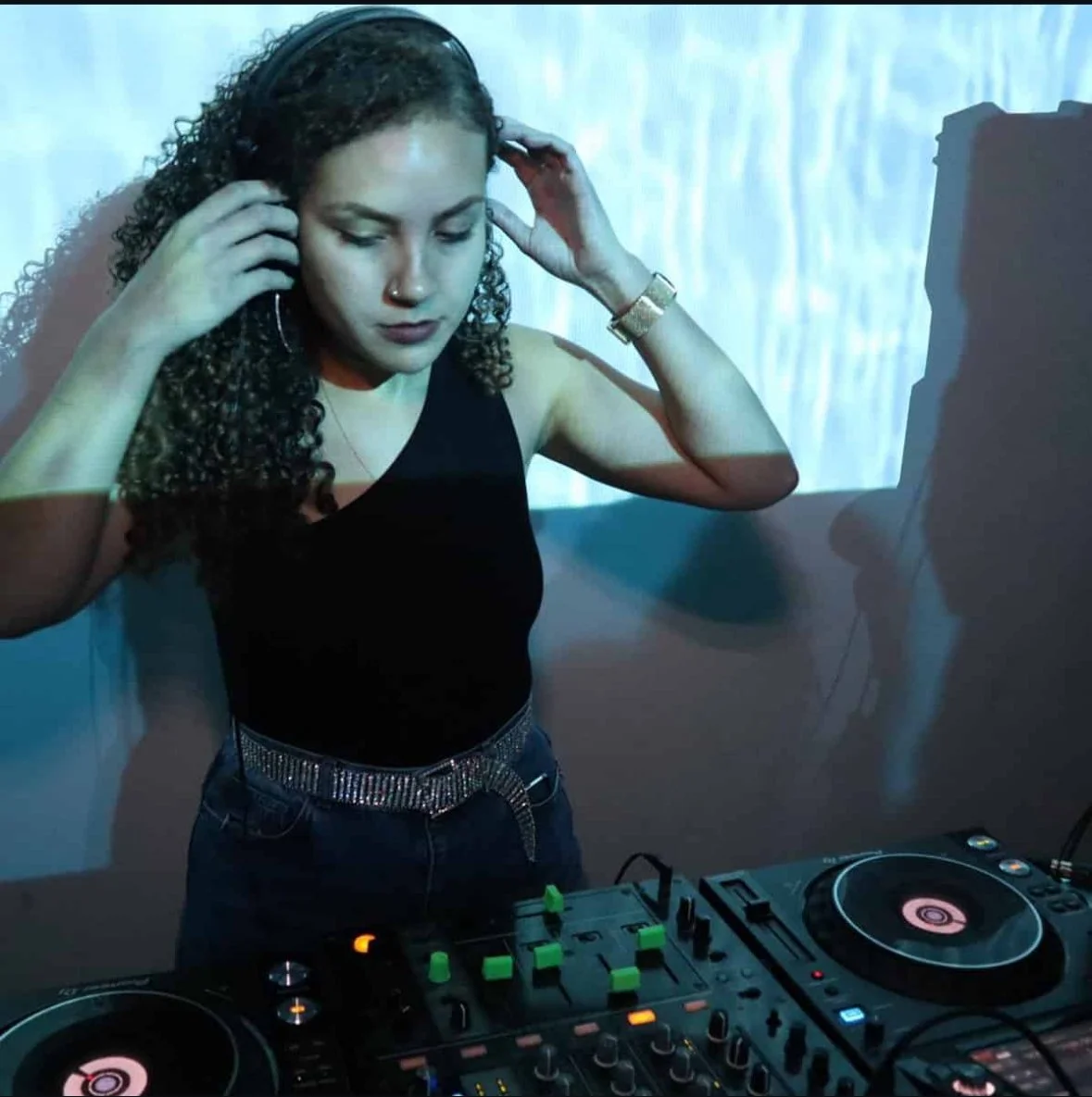 A woman with curly hair in a black sleeveless top and a sparkly belt is DJing, wearing headphones and focused on the music, with DJ equipment in front of her.