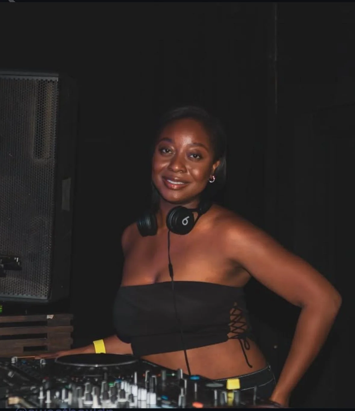 A woman wearing a black tube top with side lace-up details, standing behind a DJ console with headphones around her neck, smiling at the camera in a dark setting.