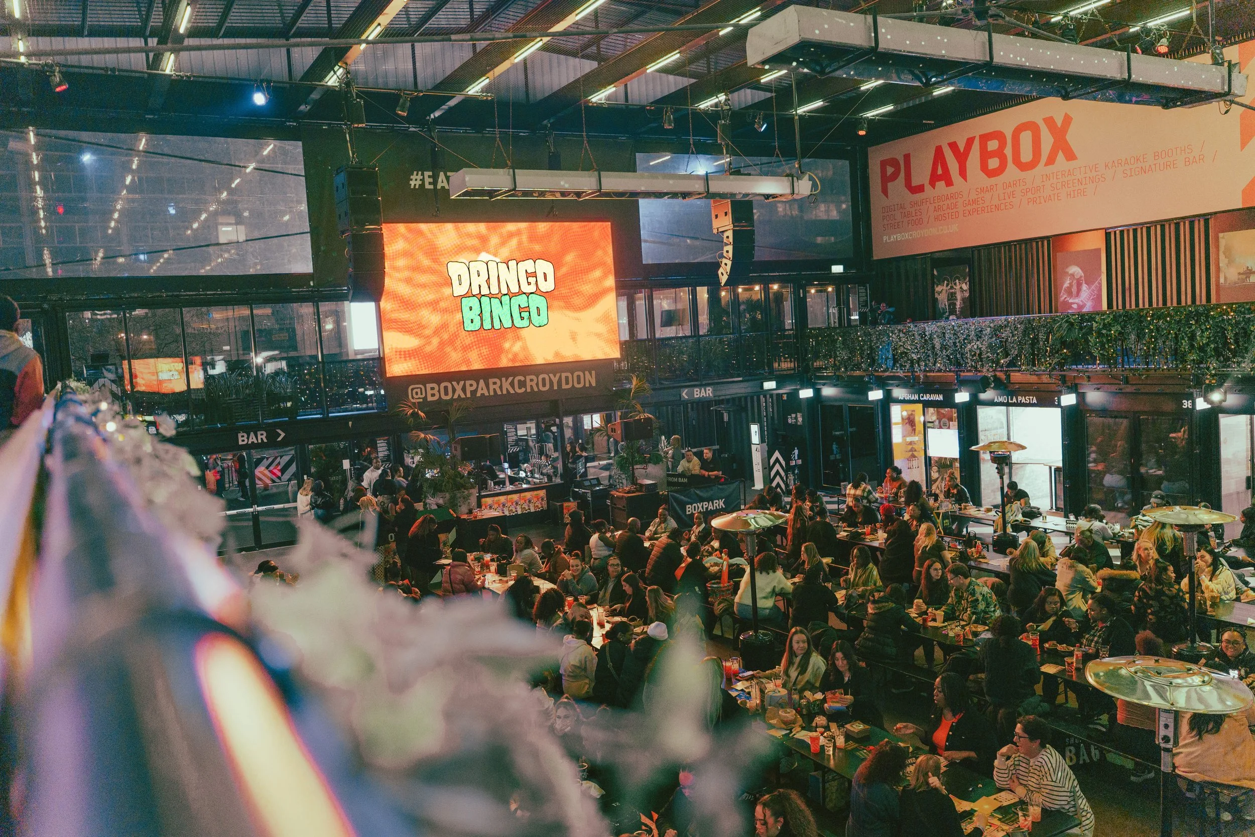 Crowd gathered in a lively indoor space with high ceilings, neon lights, bar counters, and a large digital screen displaying 'Dringo Bingo' and social media handle '@BOXPARKCROYDON'. Red 'PLAYBOX' sign and various advertising banners are visible. The area appears to be a venue for entertainment and social events.