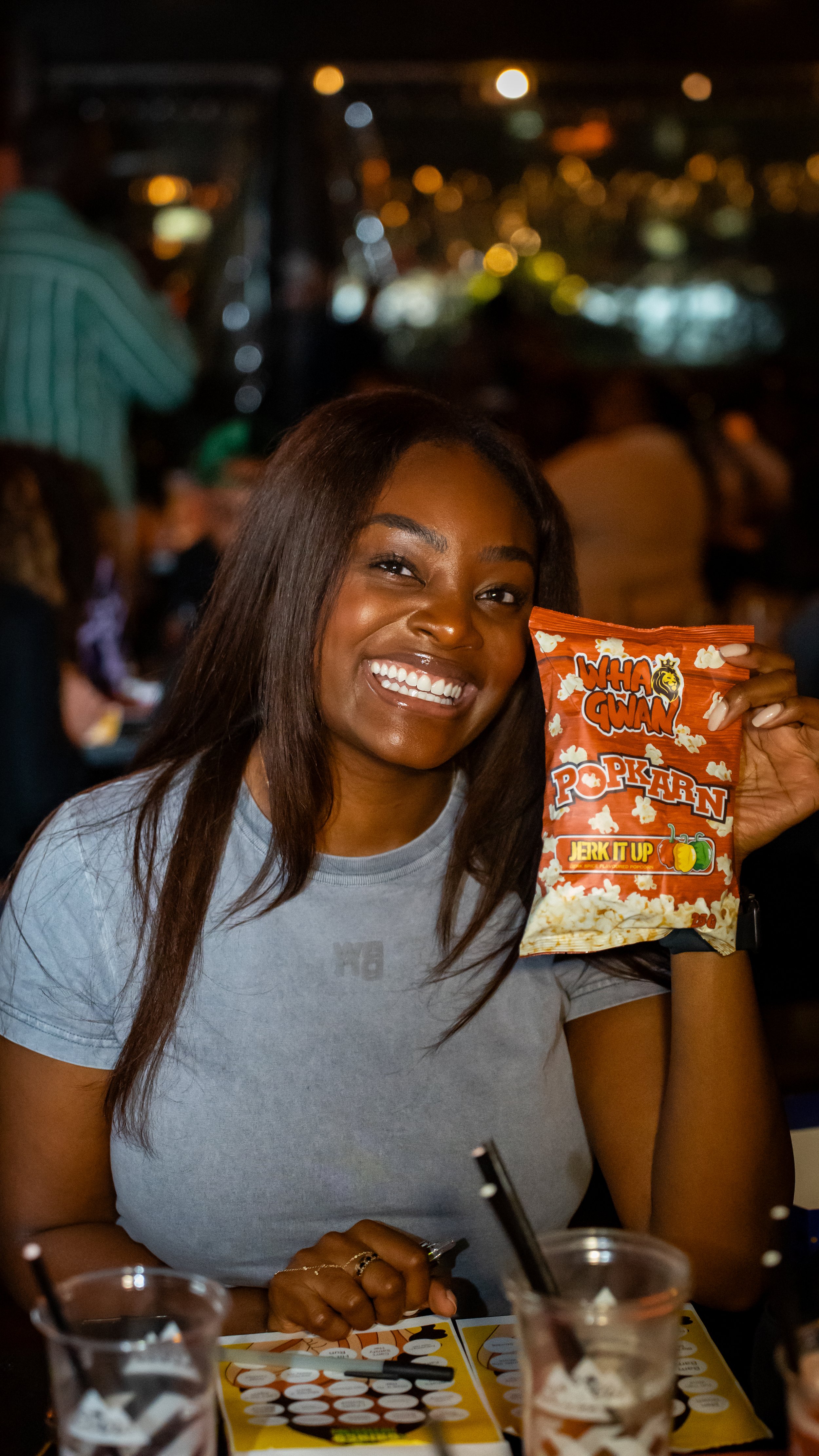 A woman with long dark hair smiling and holding a bag of popcorn at an outdoor night event with city lights in the background.