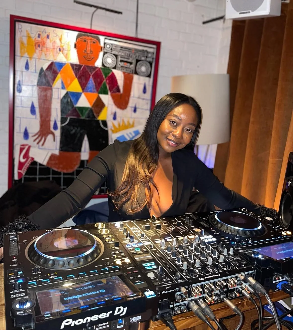 A woman is DJing at a party, standing behind a DJ console with turntables and mixers, smiling at the camera. Behind her, there is a colorful, abstract art painting on a white brick wall.
