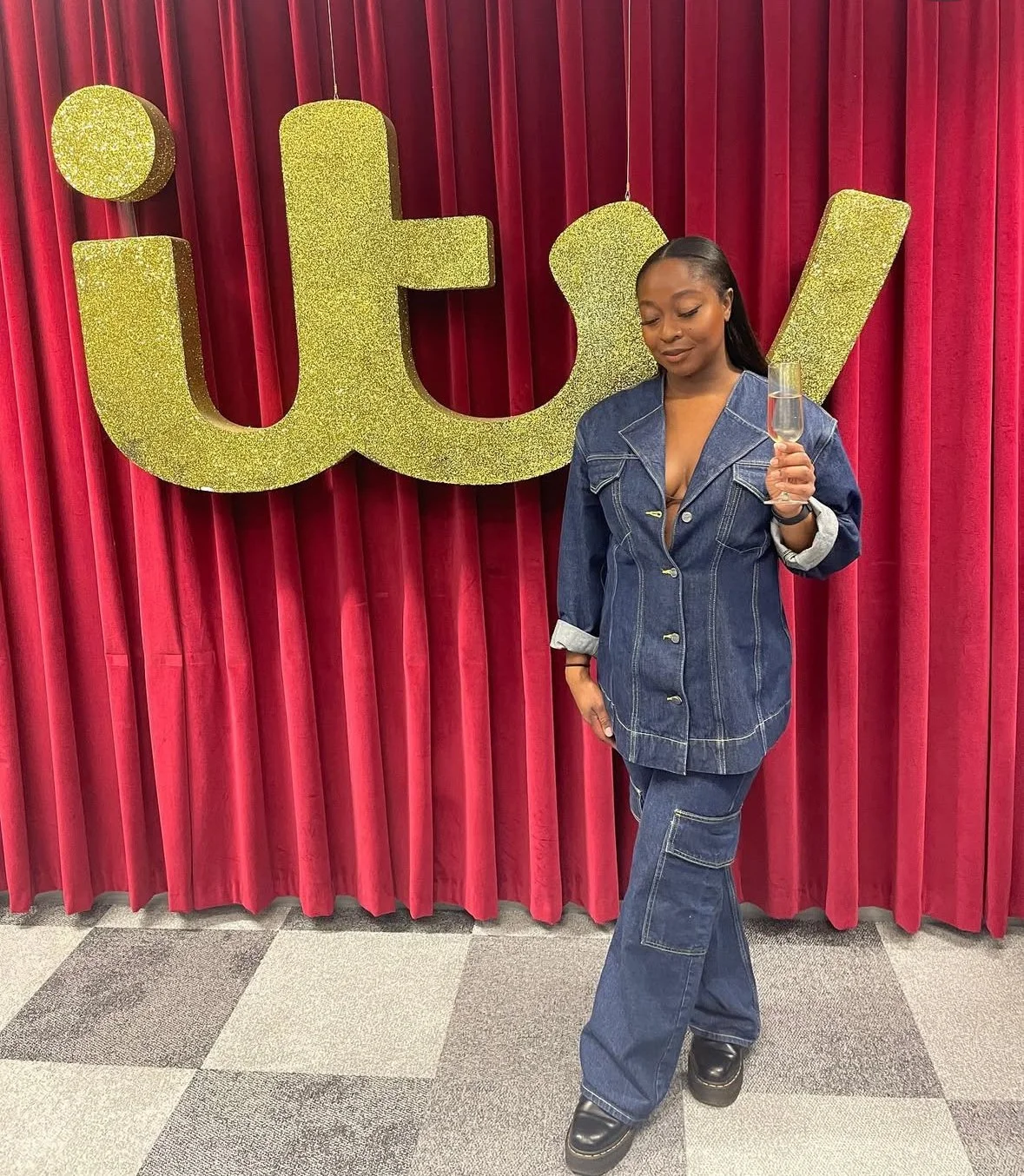 Woman in denim outfit holding a glass of champagne standing in front of red curtains with large gold glitter letters spelling 'it'