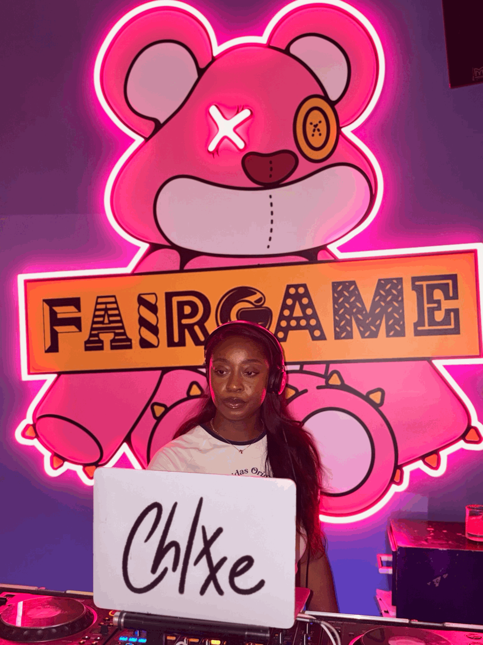 A woman wearing headphones standing in front of a neon pink and purple background featuring a cartoon bear with a sign that says 'FAIRGAME' while holding a whiteboard with the word 'Chixe' written on it.