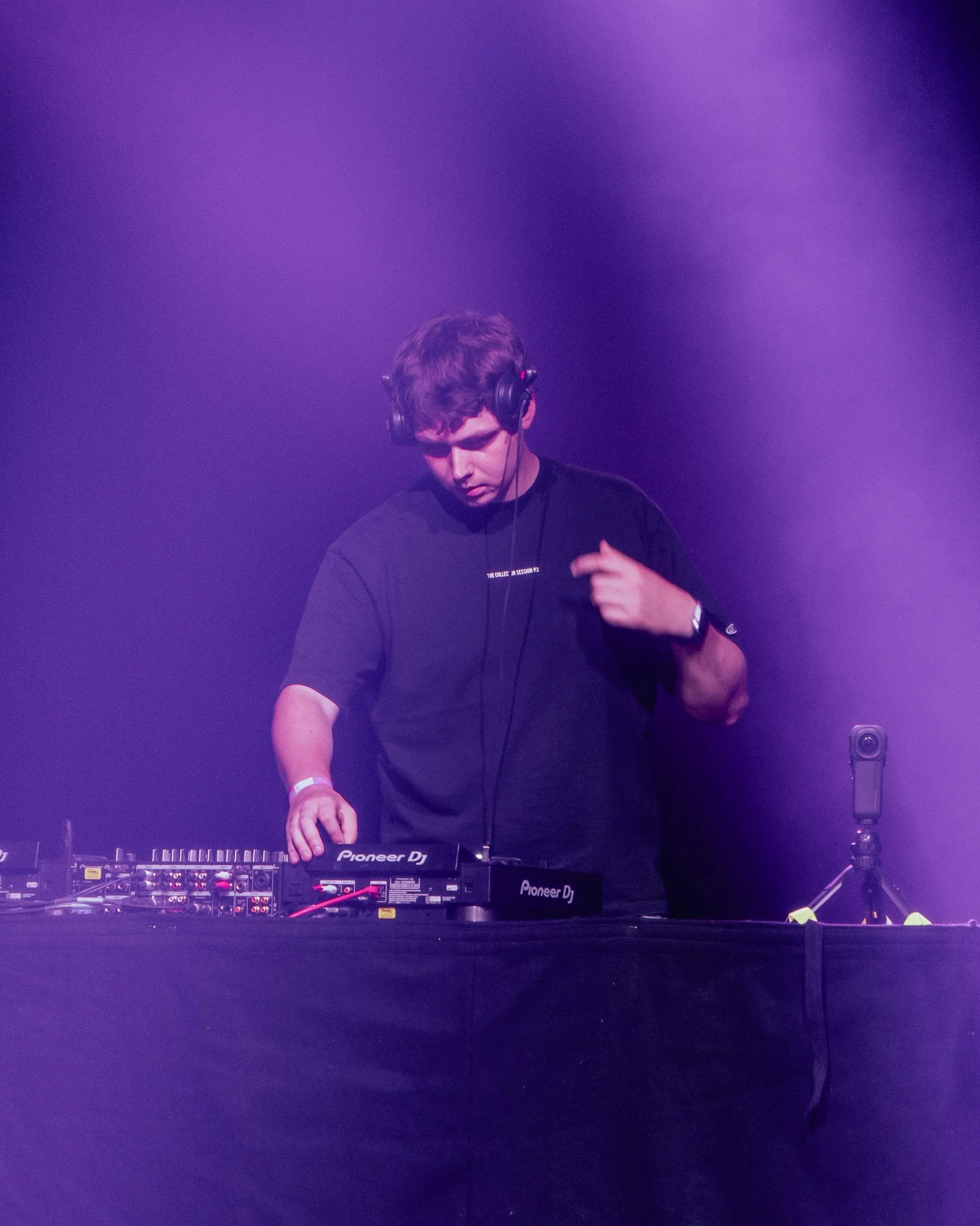 A young man DJing at a music event, wearing headphones and adjusting equipment on a table with purple stage lighting.