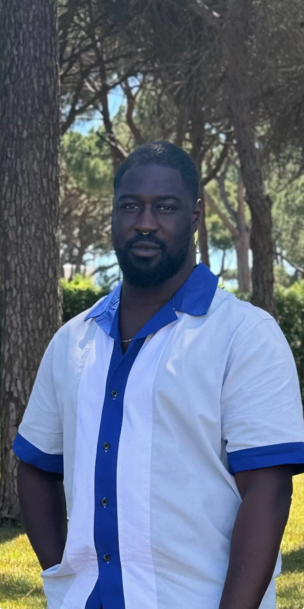 A man with dark skin, short black hair, and a beard, wearing a white and blue button-up shirt, stands outdoors near trees and greenery on a sunny day.