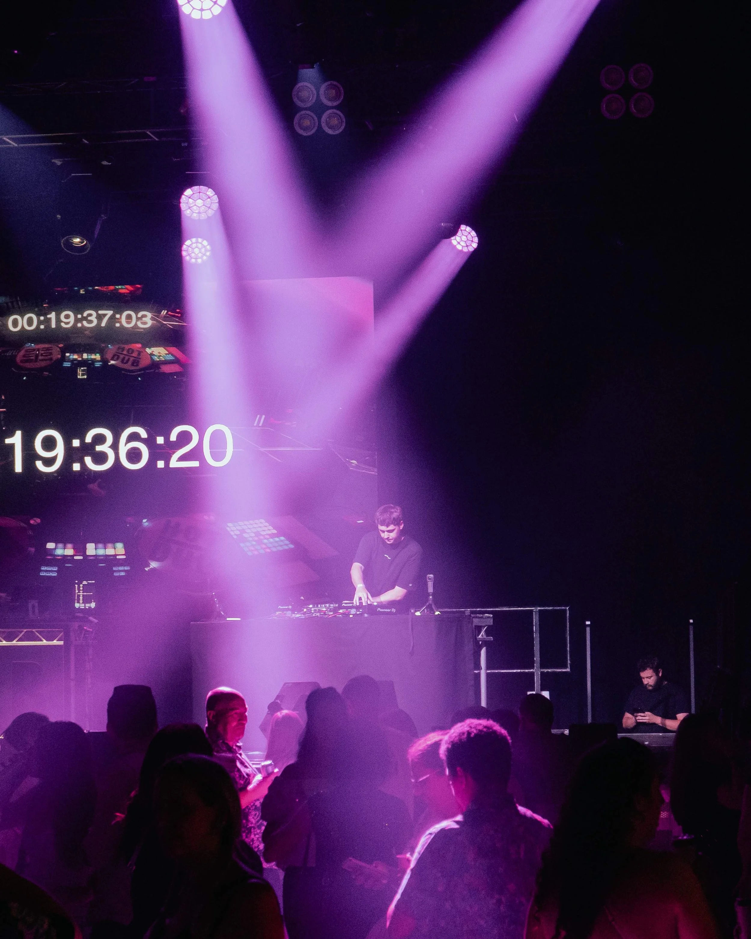 A DJ performing on stage with purple lighting effects, with a crowd of people dancing in front. A large screen displaying a timer is visible behind the DJ.