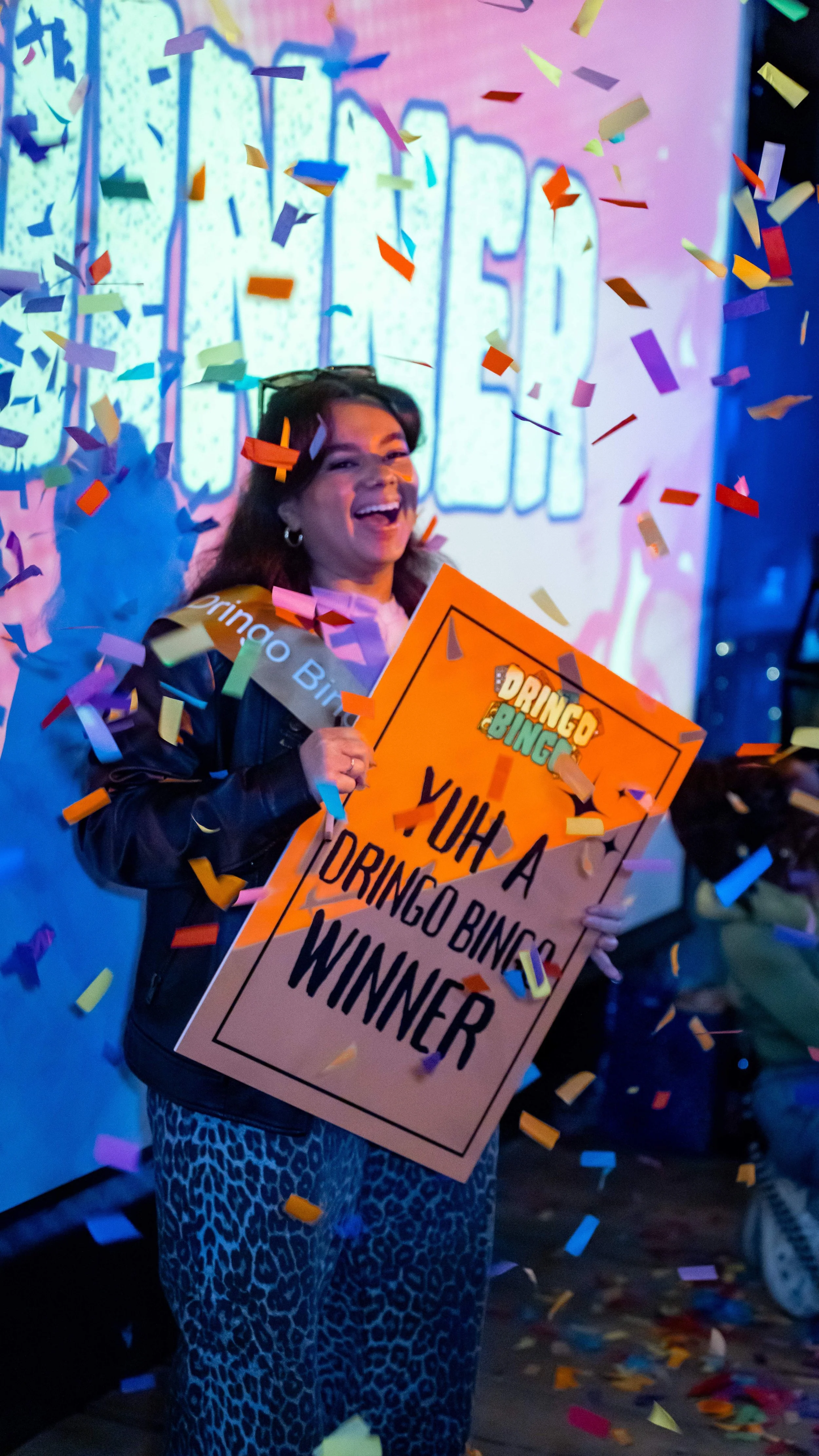 A woman celebrating a win at a Bingo event, holding a sign that says "YUH A DINGO BINGO WINNER," surrounded by falling colorful confetti, with a broad smile and festive atmosphere.