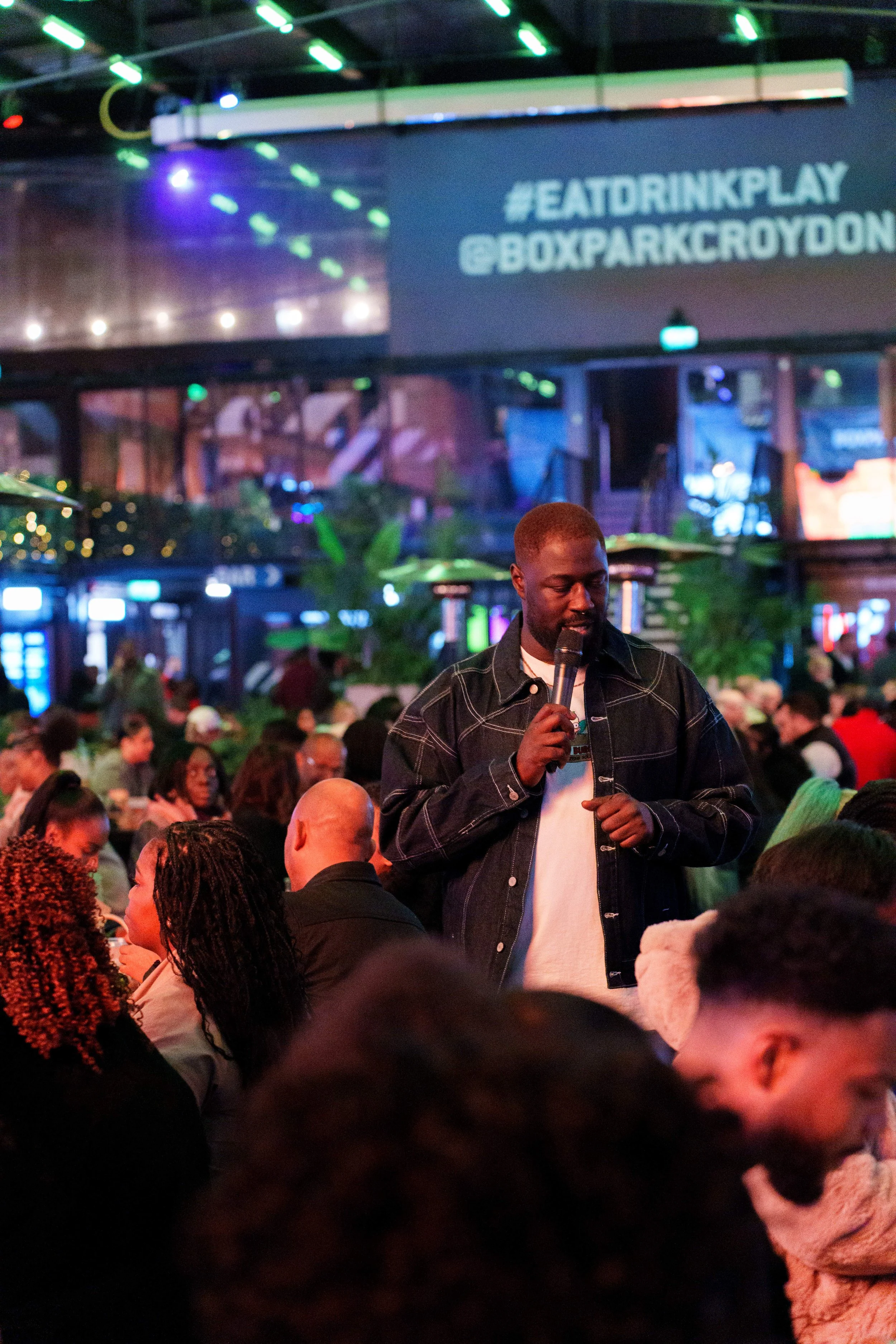 A man holding a microphone speaking to a crowd in a dimly lit, indoor event space with neon lights and a large screen showing the hashtag #EATDRINKPLAY and the location @BOXPARKCROYDON.