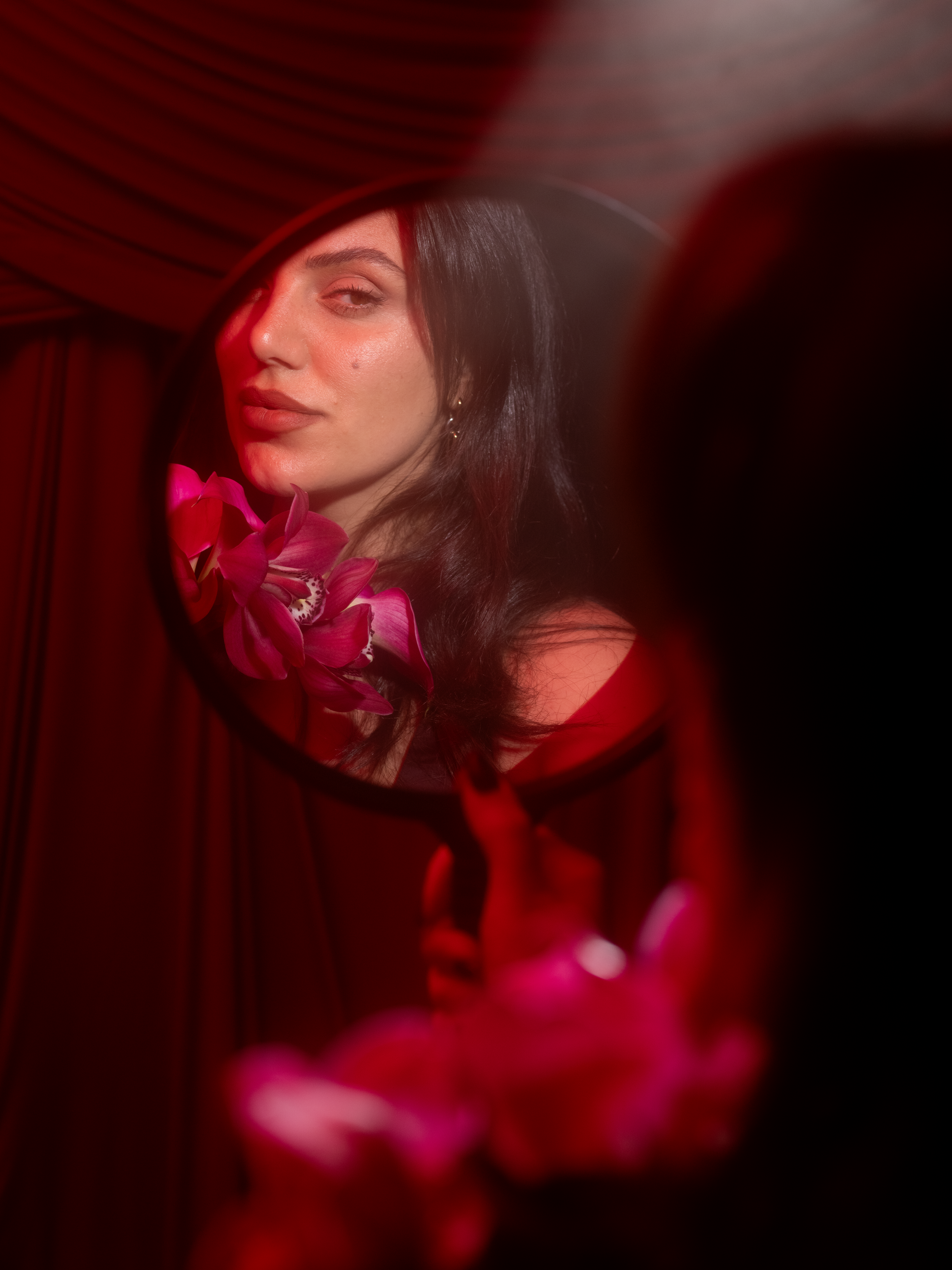 A woman with dark hair looking into a mirror, surrounded by pink orchids with warm red lighting.