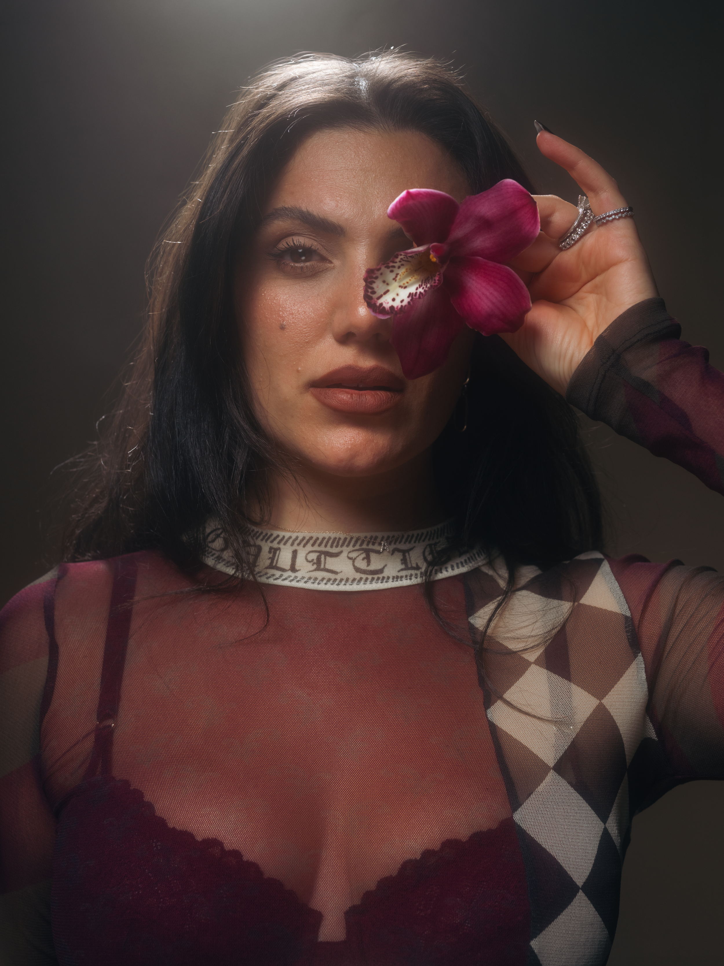A woman with dark hair and makeup holding a pink orchid flower near her face, wearing a patterned top with sheer sleeves.