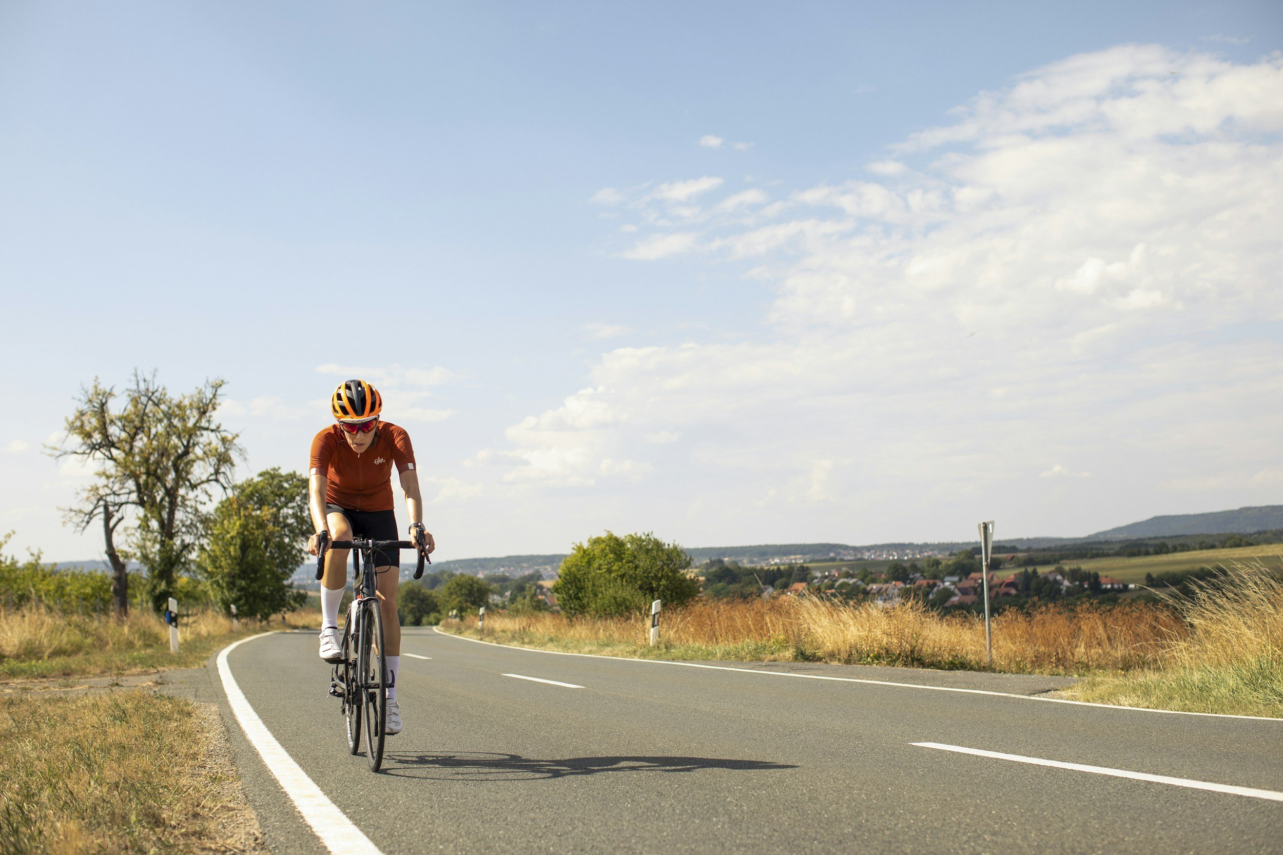 Ein Radfahrer trägt einen Helm und eine braune Trikothose, fährt auf einer kurvigen Landstraße bei Sonnenlicht.