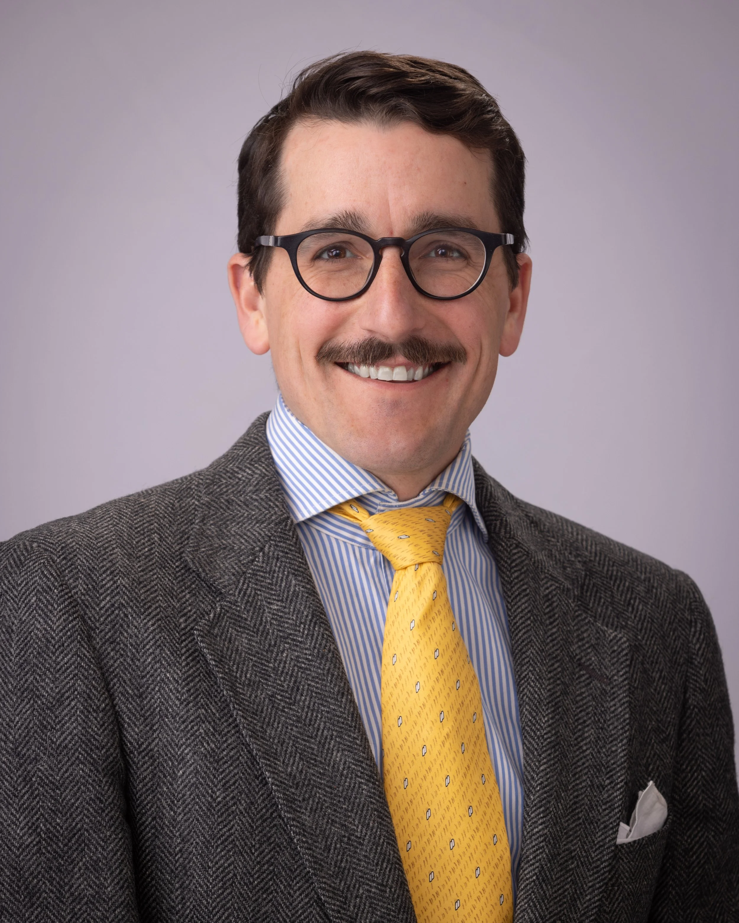 man in suit, mustache, glasses, brown hair and eyes, smiling, yellow tie.