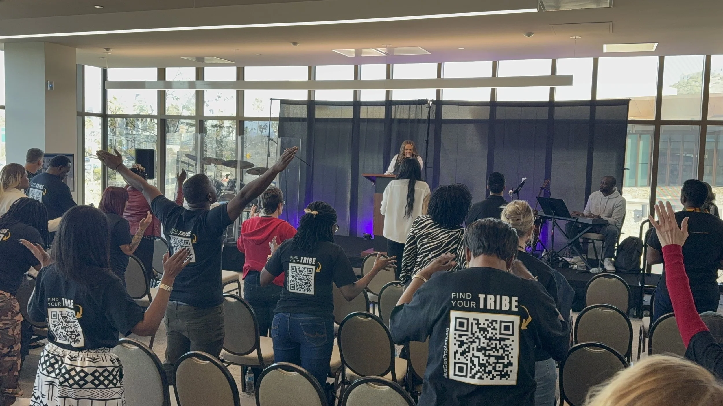People attending a conference or event in a bright, modern room with large windows. Some participants are standing with hands raised, others are clapping or watching a speaker at the front. There is a stage with a woman speaking, and musicians are pl