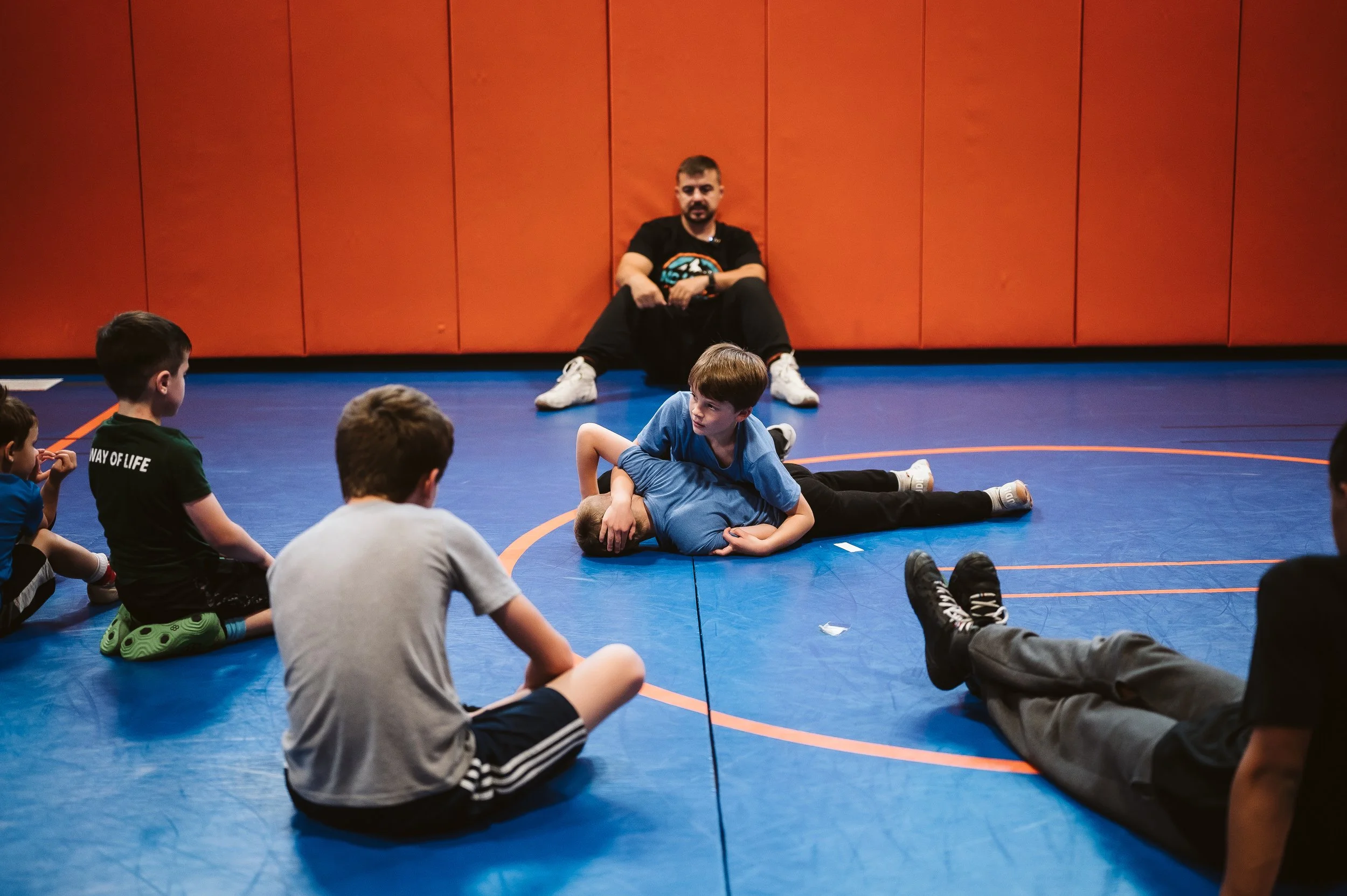Youth wrestlers practicing in a supportive environment while a coach observes, emphasizing confidence building, emotional regulation, and healthy development through wrestling.