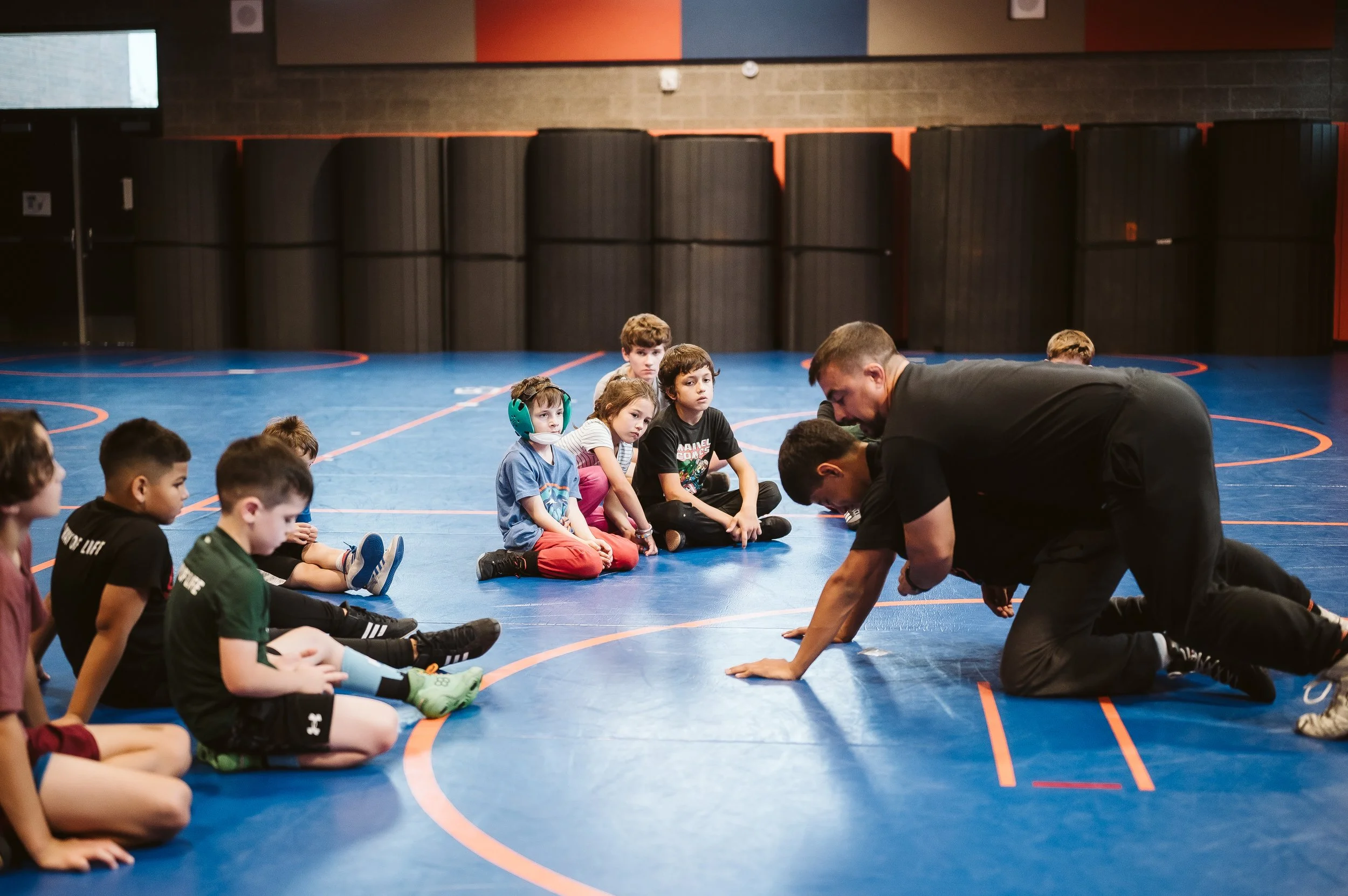 Youth wrestling coach calmly demonstrating technique to young athletes, creating a safe learning environment that builds confidence, focus, and emotional security in children’s sports training.