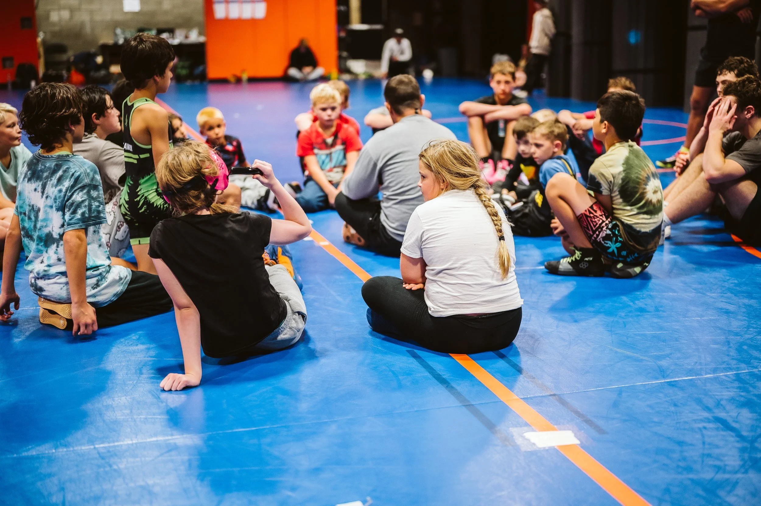 Youth wrestlers sitting in a circle on the mat during a coach-led discussion at wrestling practice, focused on mindset training and emotional regulation.