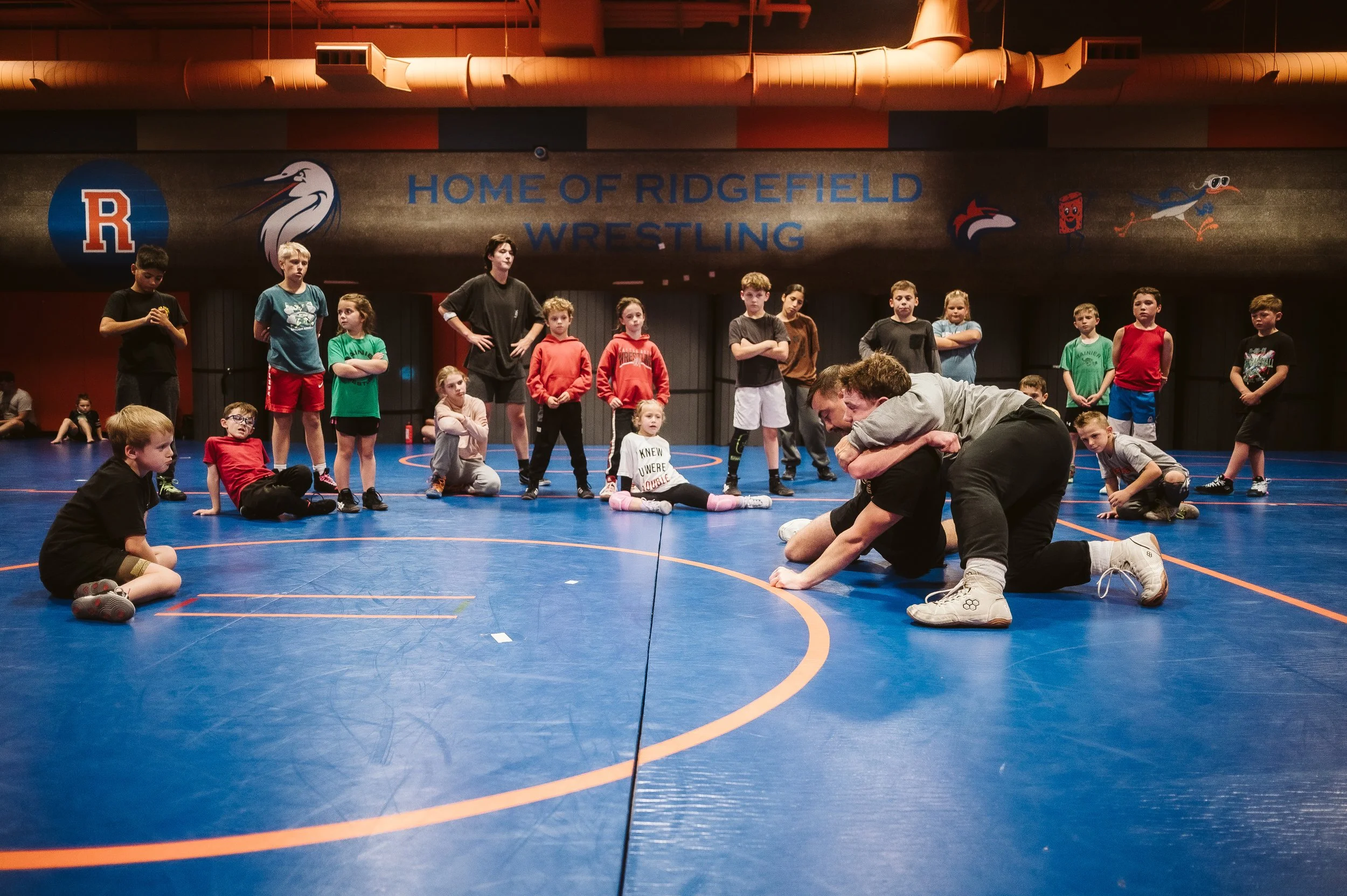 Youth wrestlers observing a coach guiding an athlete through a controlled training moment during wrestling practice, emphasizing emotional regulation and skill development.