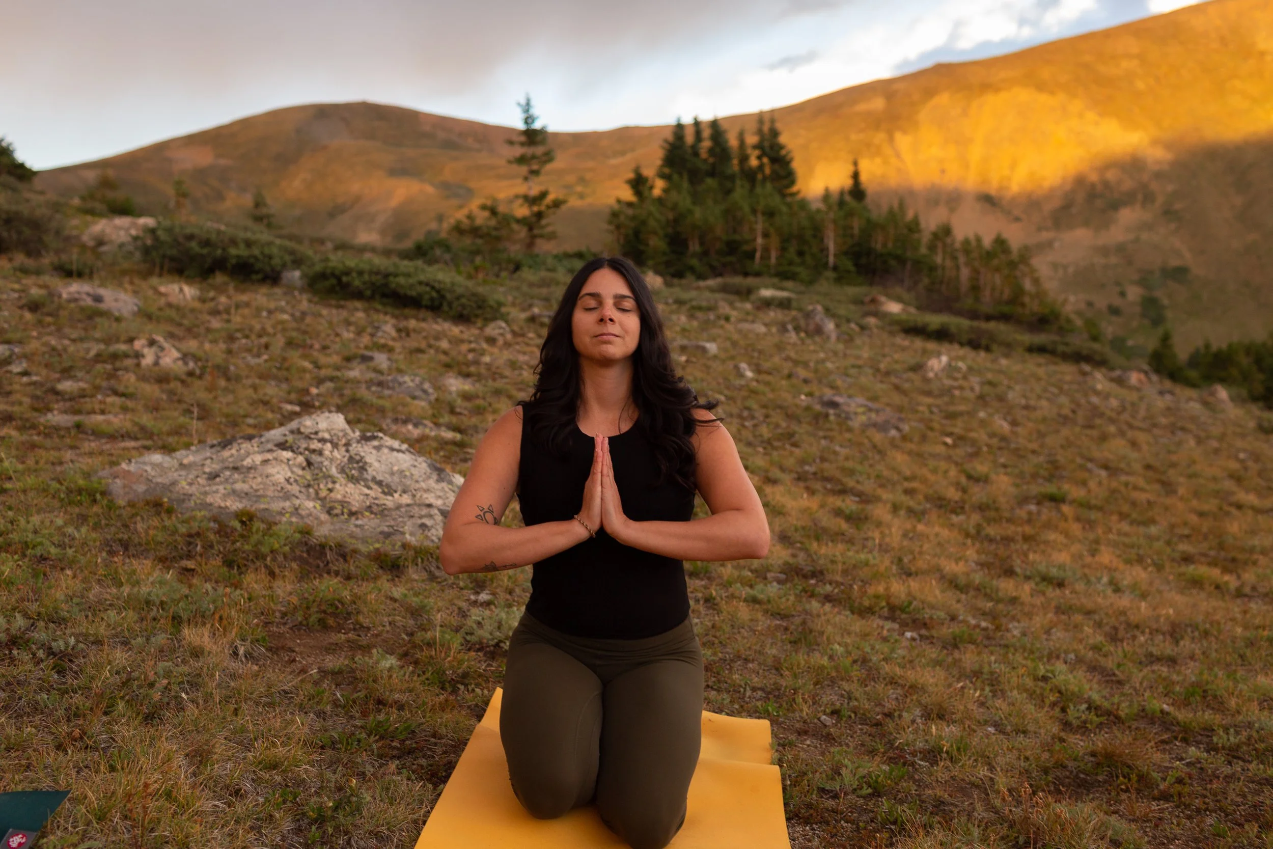Woman meditating with her hands together at her heart, mountain scene is in the background.