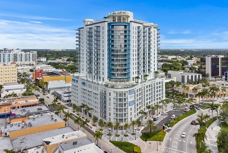 Tall modern high-rise apartment building with curved glass balconies, surrounded by palm trees in an urban area under a blue sky.
