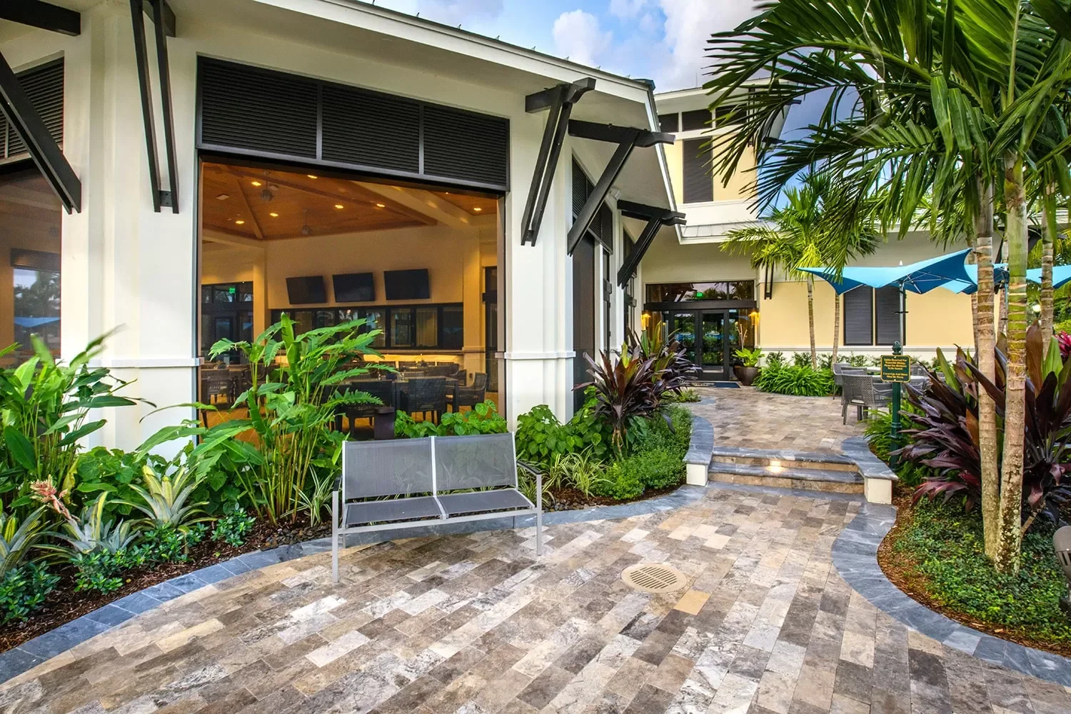 A modern outdoor area with a paved walkway, tropical plants, and seating, leading to a building with large windows and a view of the interior lounge area.