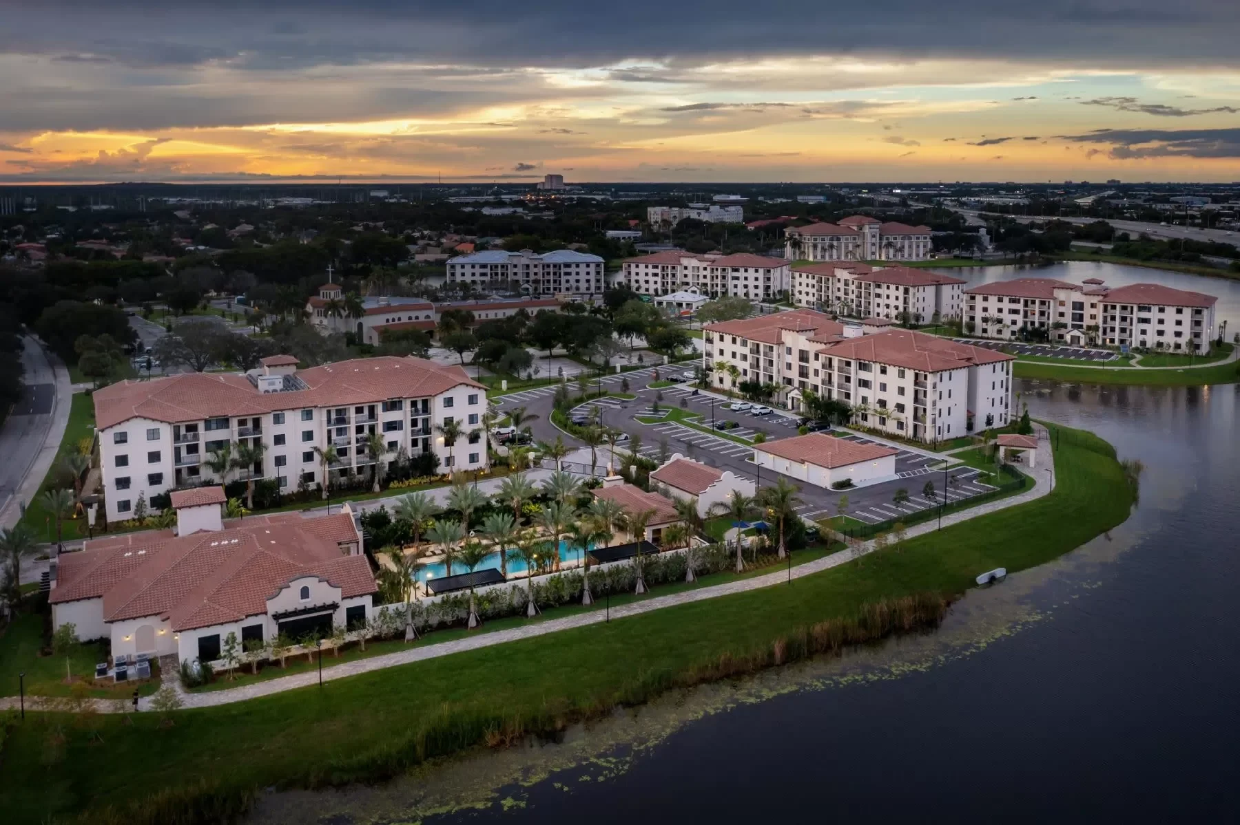Aerial view of a residential complex with multiple white buildings with red-tiled roofs, a swimming pool surrounded by palm trees, parking lots, and a waterway, during sunset.