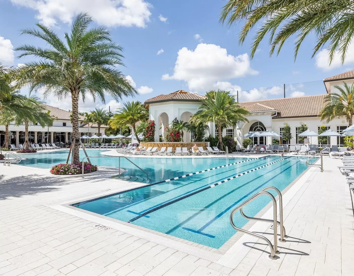 A resort swimming pool with palm trees, lounge chairs, umbrellas under a partly cloudy sky.