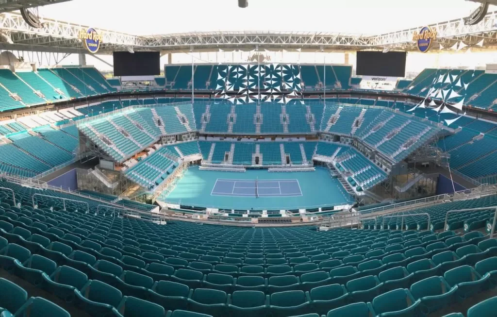 Empty tennis stadium with teal seats and a blue tennis court in the center, decorated with bunting and signage for Hard Rock and Verizon.