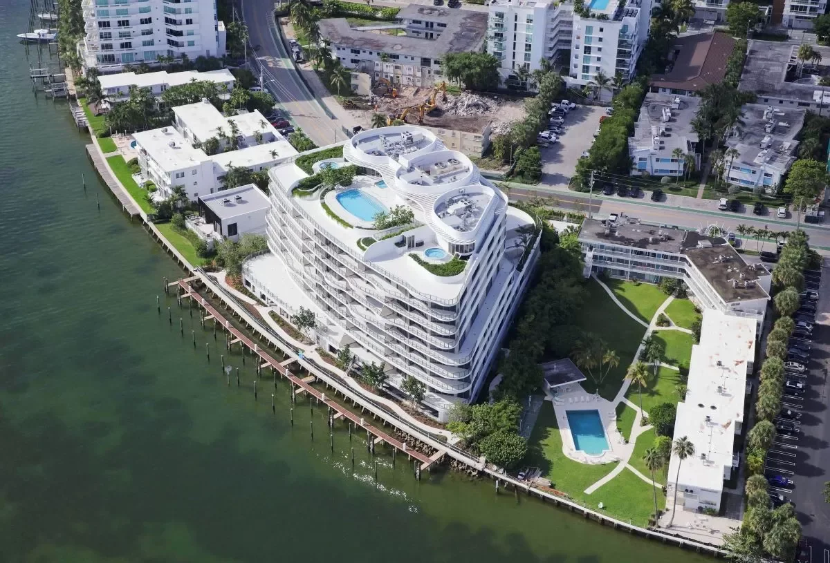 Aerial view of a modern waterfront residential complex with a large rooftop swimming pool, smaller pools, and lush green landscaping. The building is surrounded by water on one side and neighboring buildings and streets on the other.