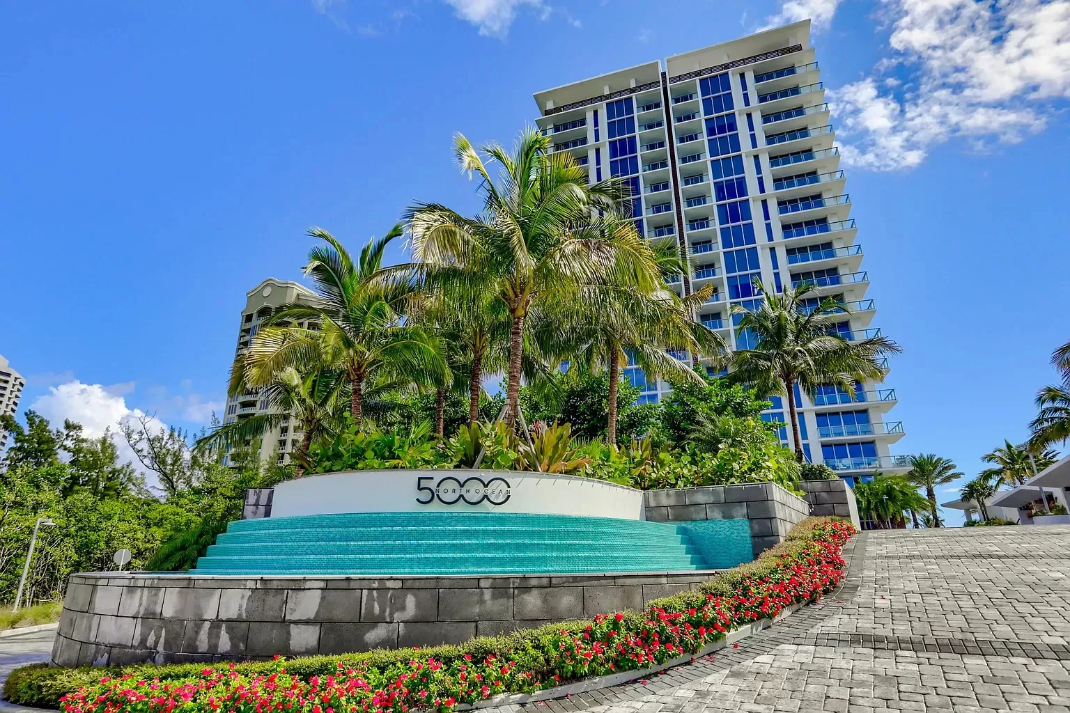 Tall modern building with glass balconies, surrounded by palm trees and colorful flower beds, under a blue sky.
