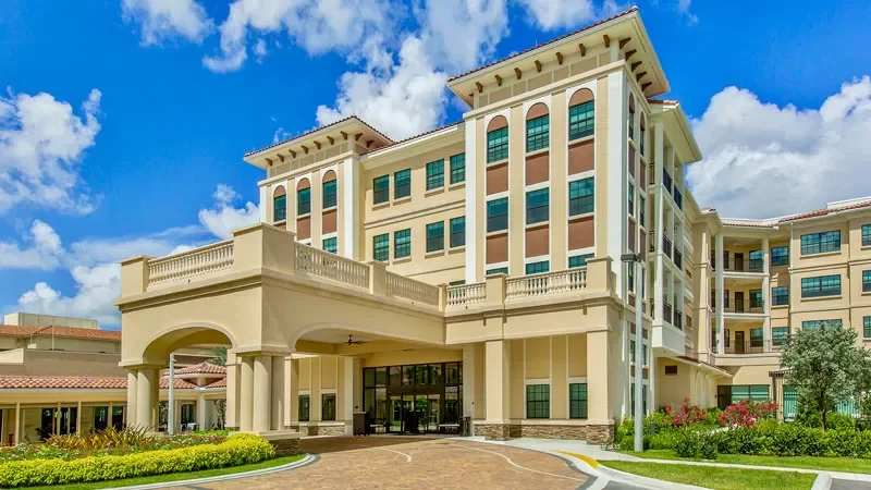A large modern apartment building with beige walls and numerous windows, featuring a covered entrance with columns, surrounded by landscaped grass and bushes, against a bright blue sky with scattered clouds.