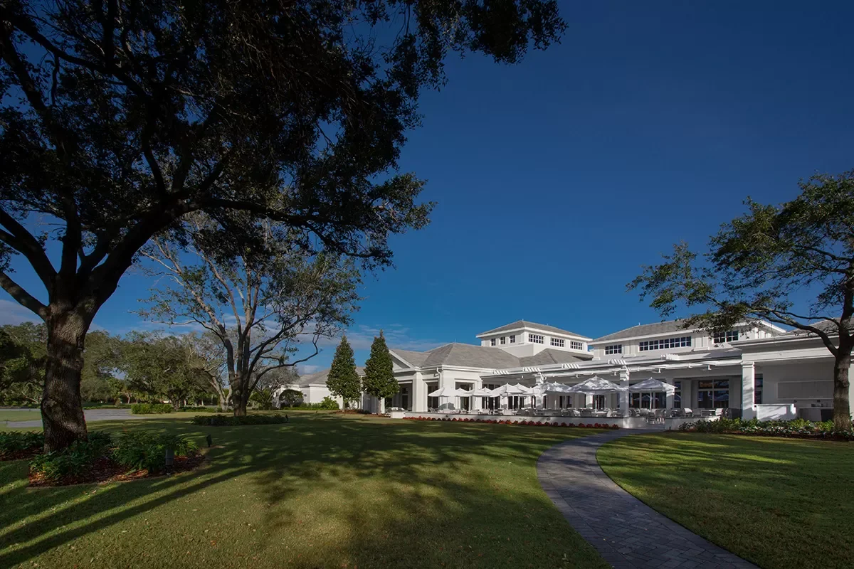 A large white building with outdoor seating and umbrellas, surrounded by a well-maintained lawn and trees, under a clear blue sky.