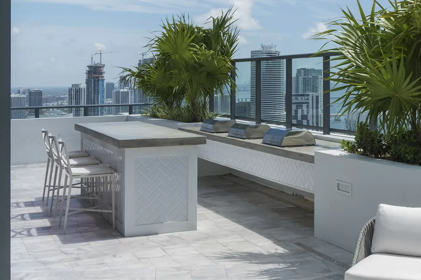 Rooftop terrace with white tile flooring, a tall dining table with four white chairs, and a built-in barbecue grill area. Large potted plants and city skyline view in the background.