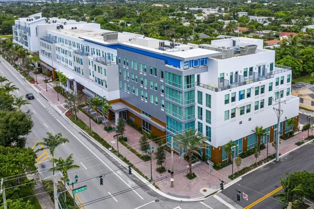 An aerial view of a modern multi-story building with white and blue exterior, surrounded by palm trees and situated along a city street.
