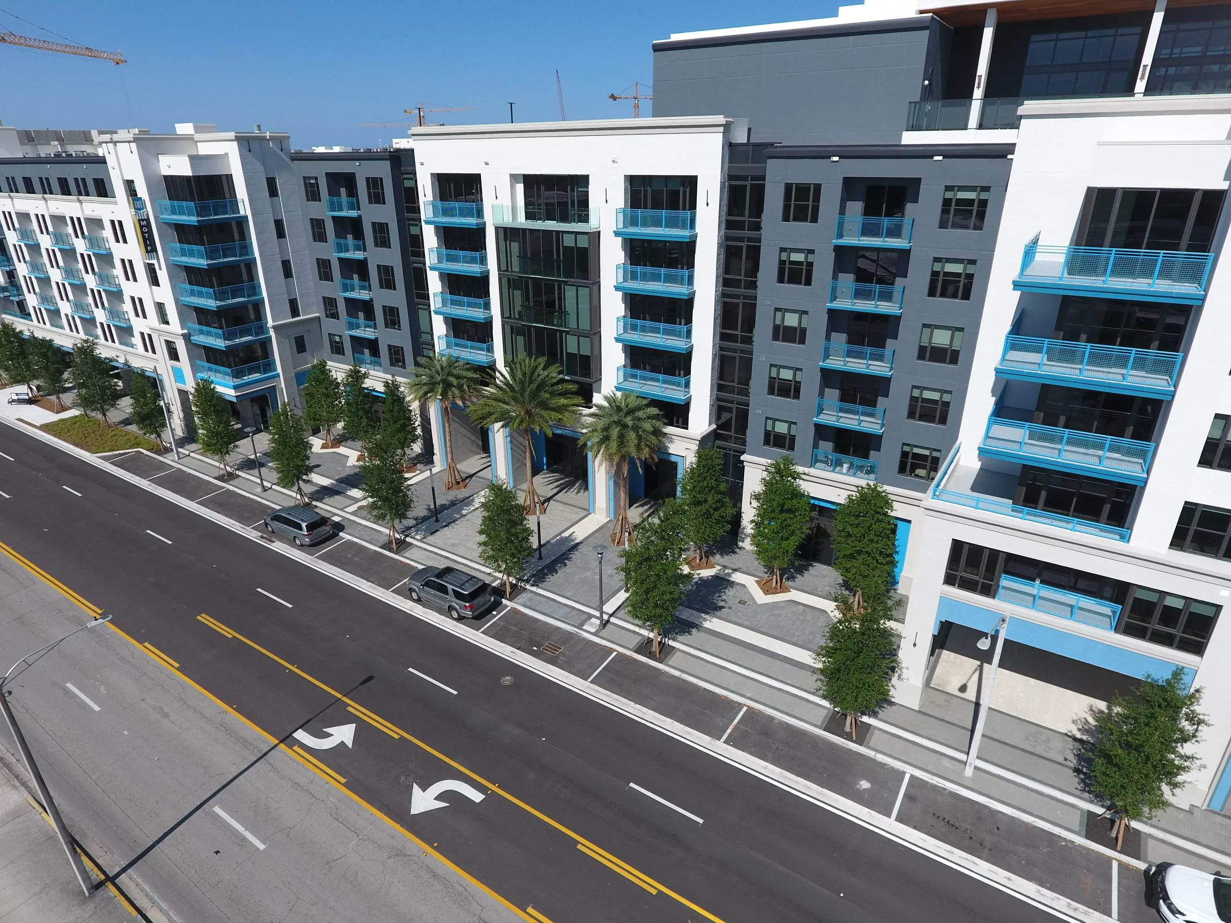 Aerial view of a modern residential apartment complex with white and gray buildings, blue balconies, palm trees lining the sidewalk, and vehicles parked along the street.