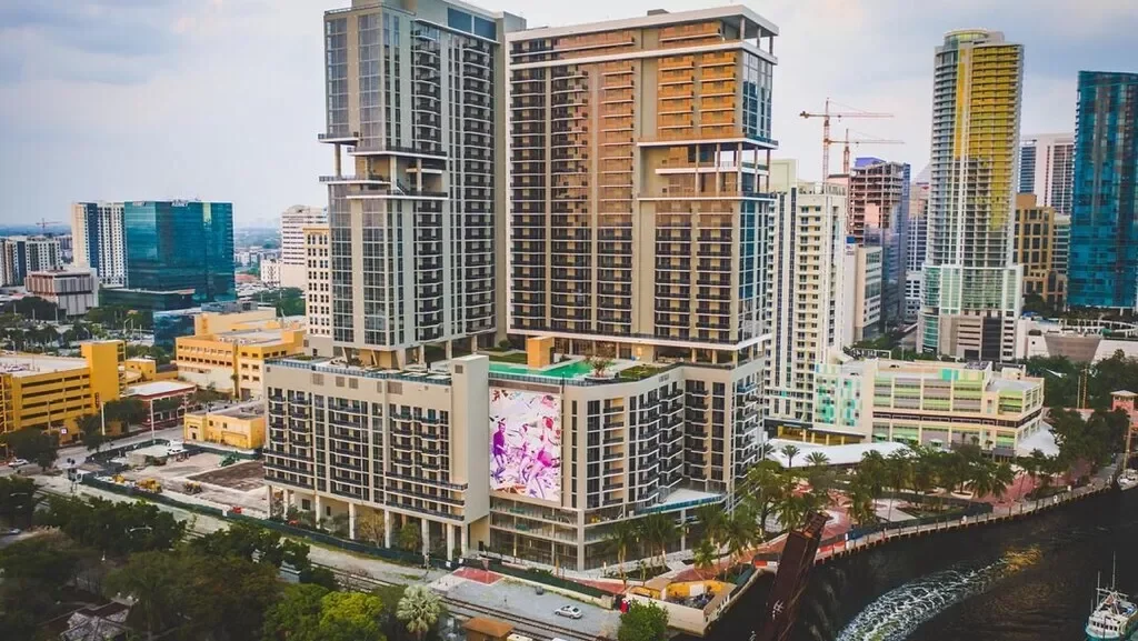 A cityscape with high-rise buildings, including a large modern residential or hotel complex with digital billboards, surrounded by additional skyscrapers, construction cranes, and a waterway with trees along the banks.