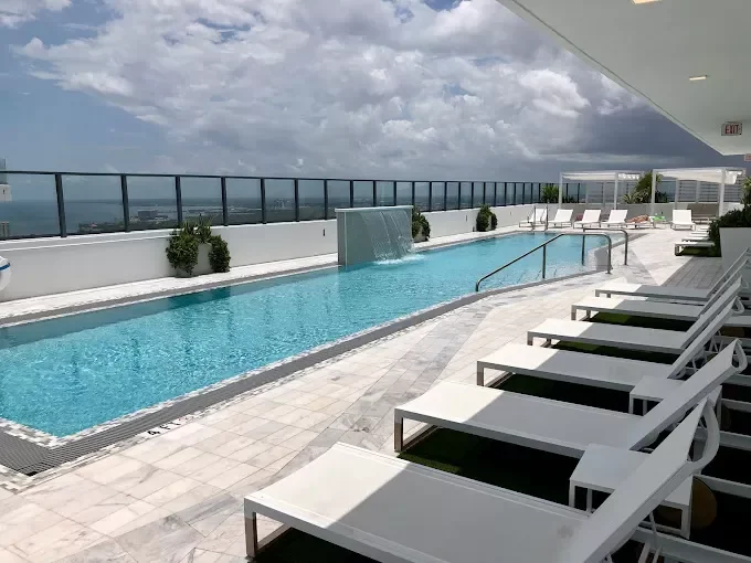 Rooftop pool with lounge chairs, glass railing, and city view with cloudy sky.
