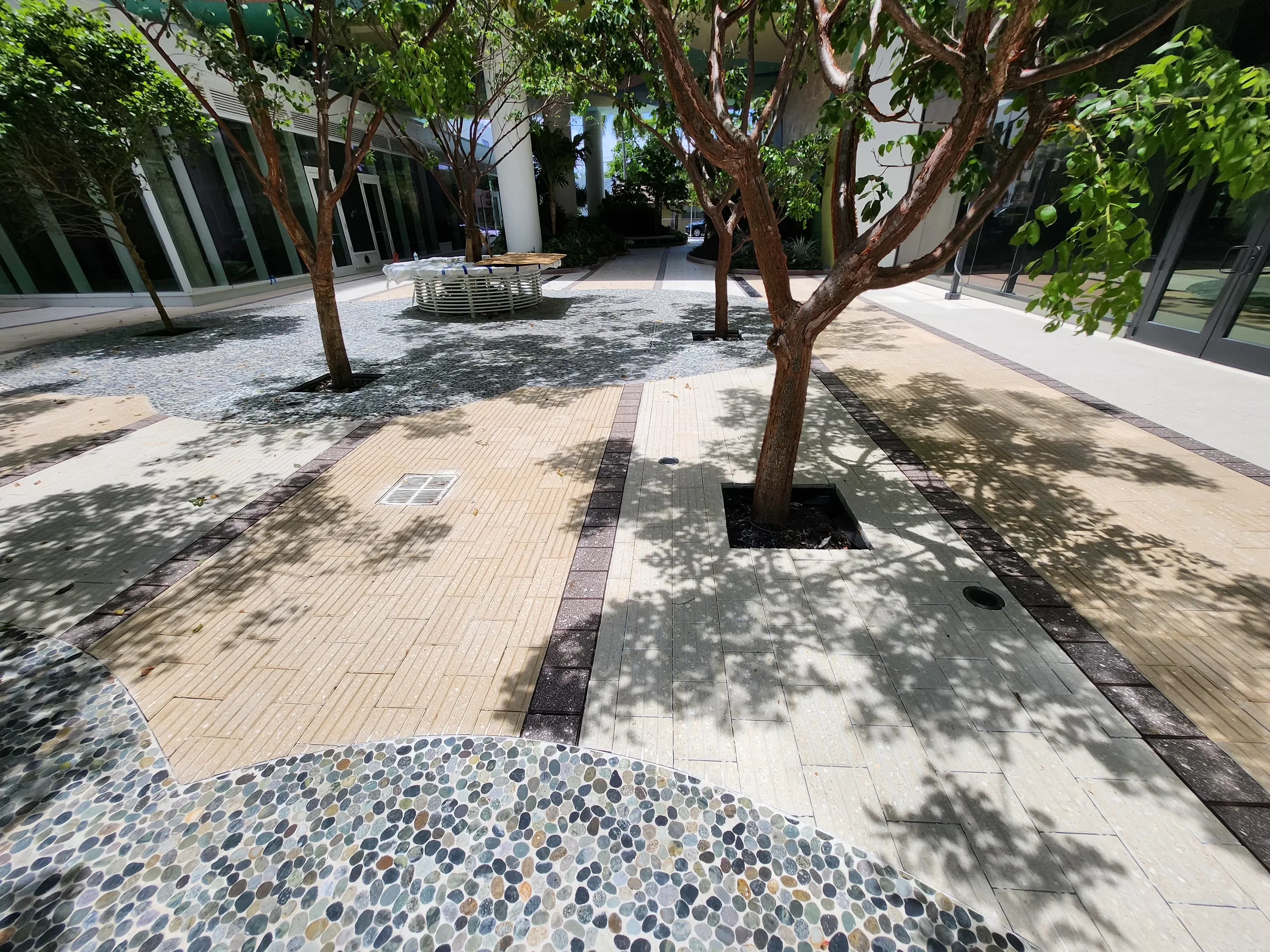A courtyard with trees providing shade, paved walkways, and modern glass buildings in the background. There is a round seating area or table further back, and the ground is covered with small pebble mosaics and tiles.