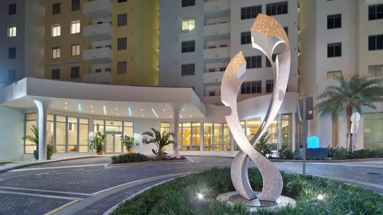 Modern hotel entrance with a metal sculpture in the foreground, tall palm trees, potted plants, and a multi-story building in the background at night.