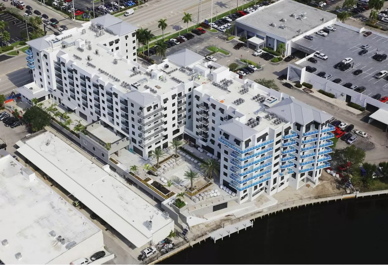 Aerial view of a white multi-story apartment complex with a waterfront, featuring balconies with blue accents. Surrounding the building are parking lots, some with cars, and a parking garage. There are also some trees, shrubs, and palm trees near the complex.
