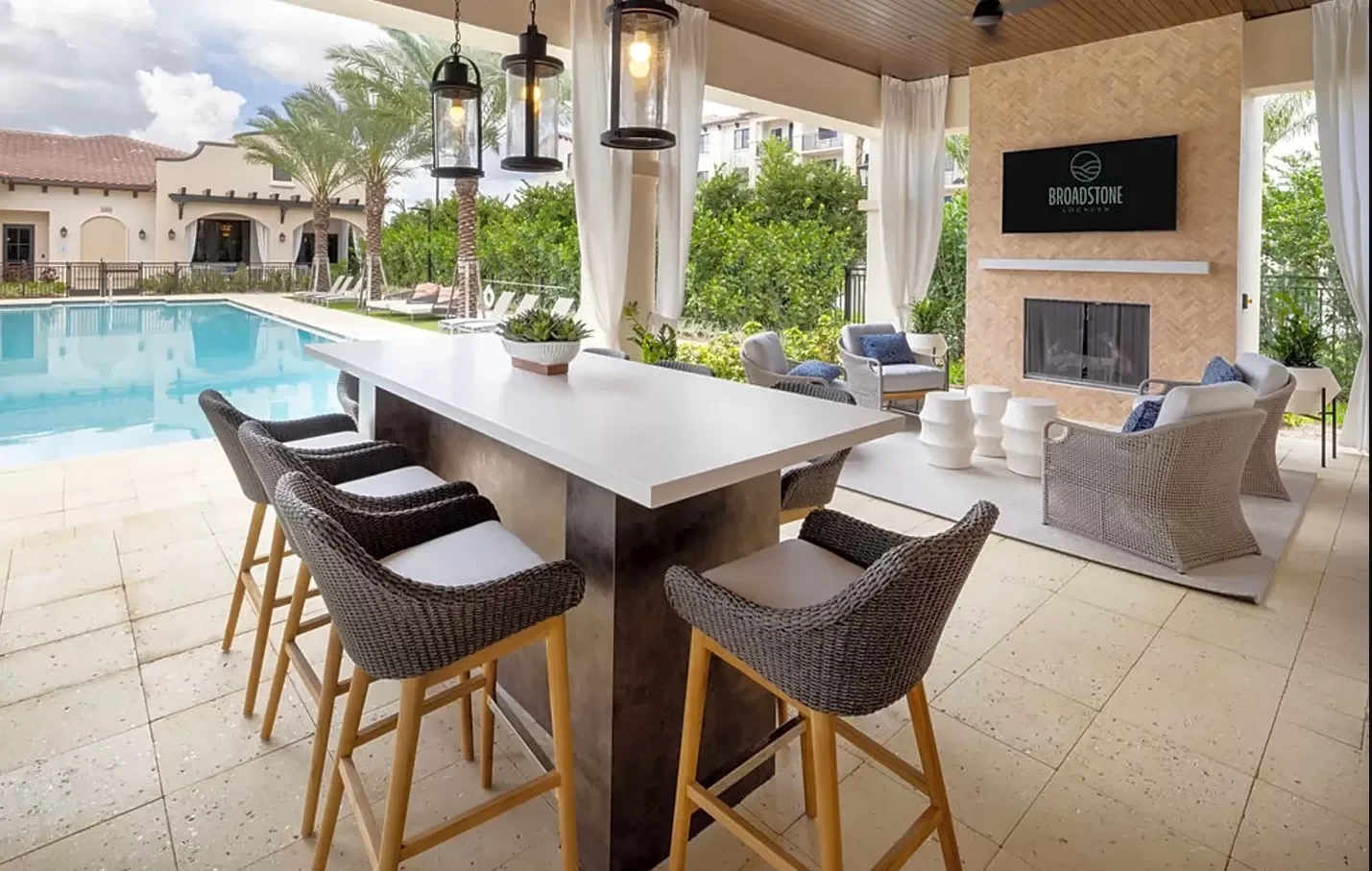 Outdoor poolside area with a white bar counter and black wicker chairs, adjacent seating area with wicker chairs and a fireplace, a screen with 'Broadstone' on it, surrounded by greenery and palm trees.